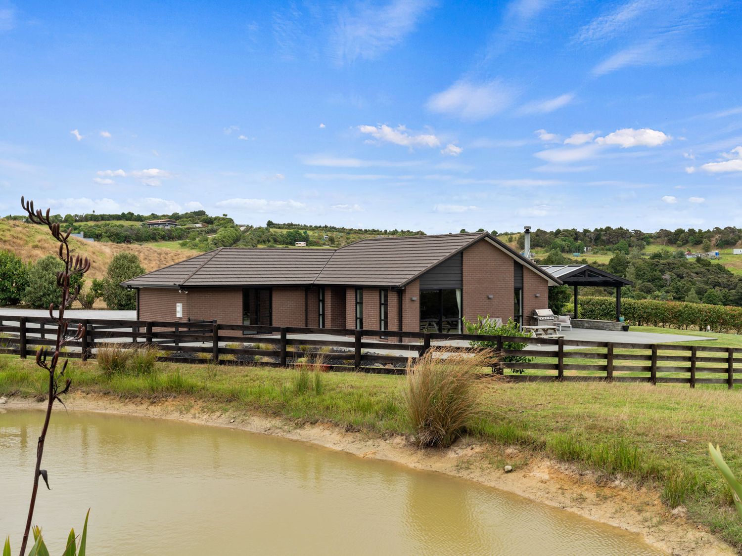 A single-story brick house with a tiled roof near a pond and wooden fence in a rural setting at Mangawhai Maketu Mangawhai