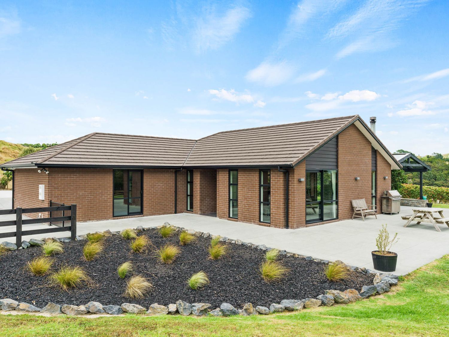 A single story brick house with a sloped roof and several windows overlooking a patio area with outdoor furniture and a garden bed at Mangawhai Maketu - Mangawhai Holiday Home in Mangawhai