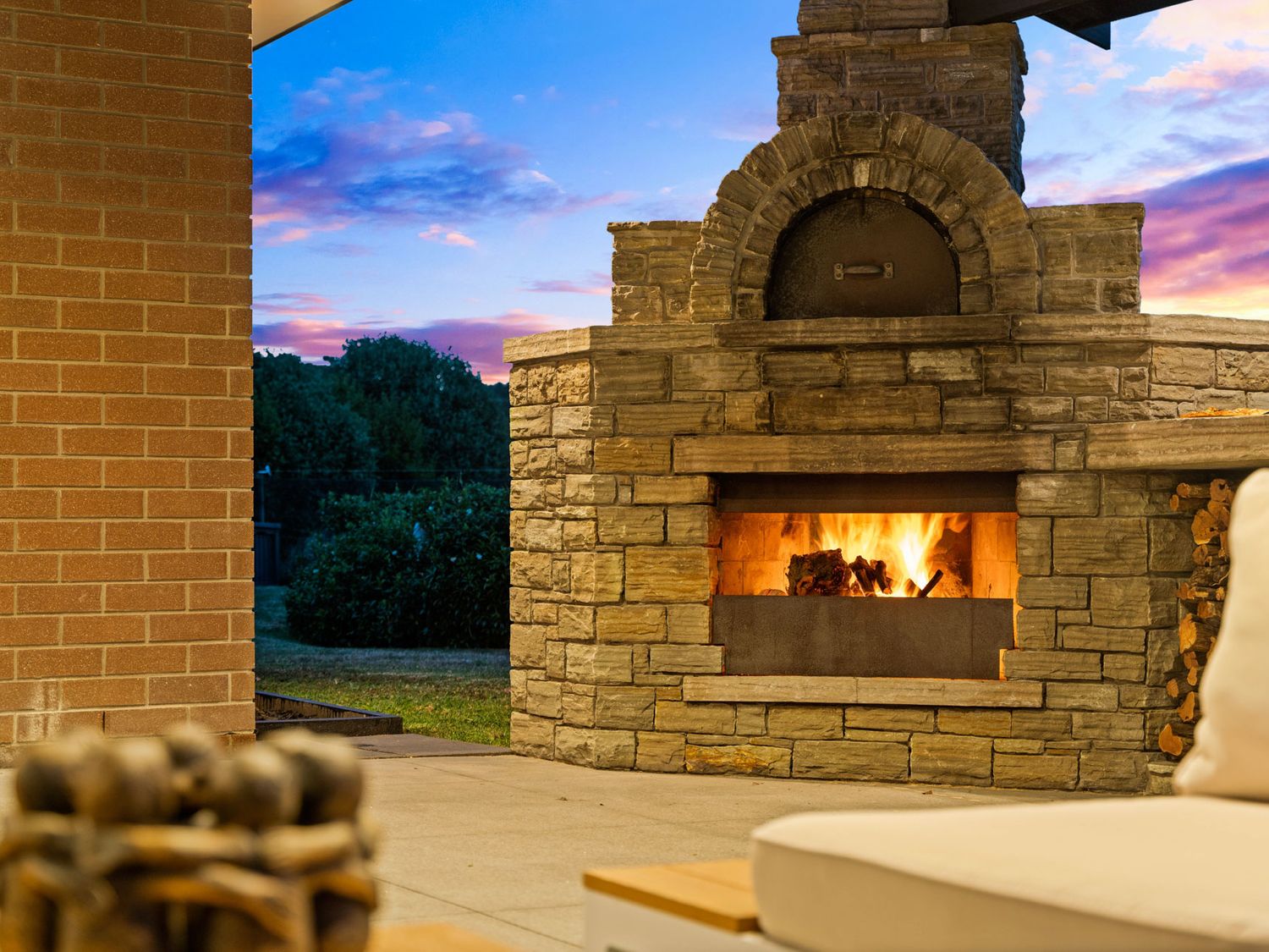 An outdoor stone fireplace with fire burning near a brick wall and furniture at Mangawhai Maketu - Mangawhai Holiday Home Mangawhai