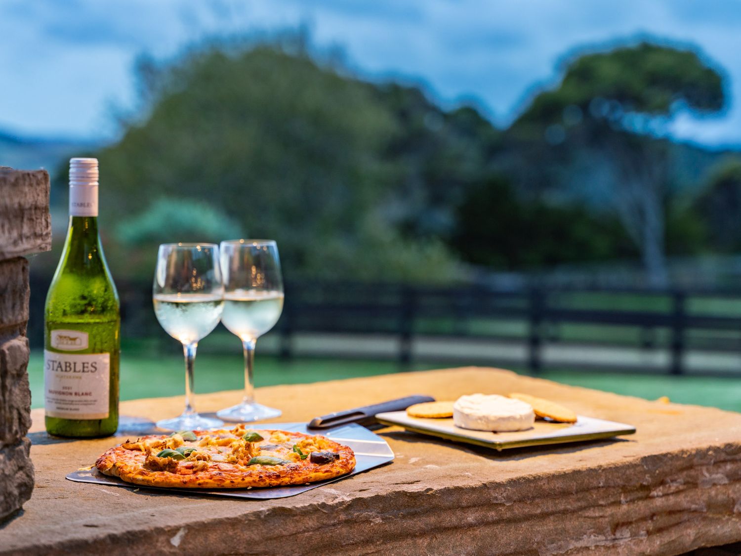 A bottle of white wine with two glasses of wine a pizza and cheese with crackers on a stone table outdoors at Mangawhai Maketu Mangawhai Holiday Home