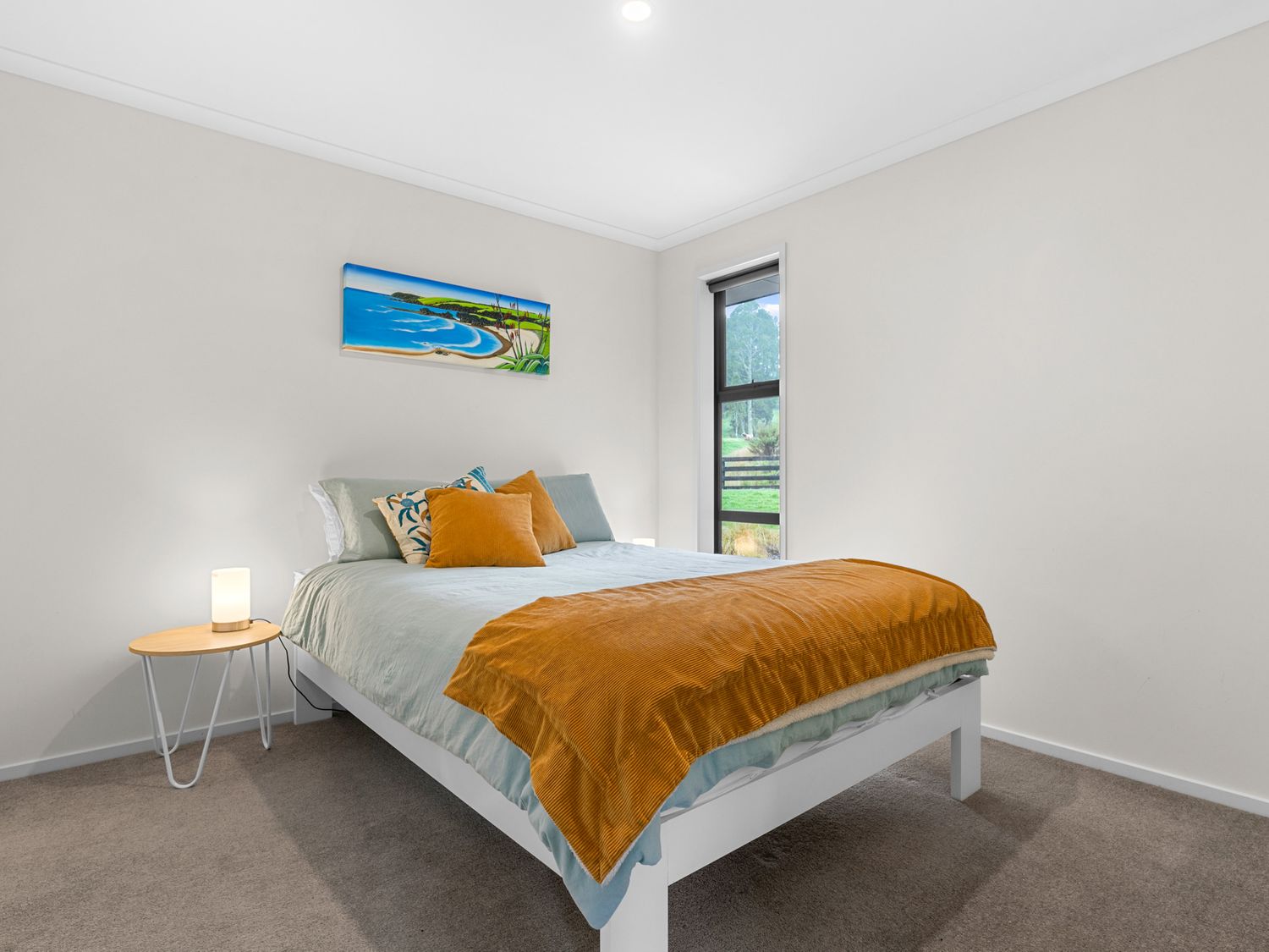 A bedroom with a bed with orange pillows and blanket a bedside table with a lamp and a window with a view at Mangawhai Maketu Mangawhai Holiday Home