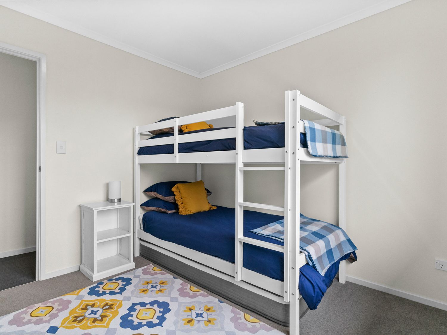 A bedroom with a white bunk bed with blue bedding a white bedside table with a lamp and a patterned rug on the floor at Mangawhai Maketu Mangawhai