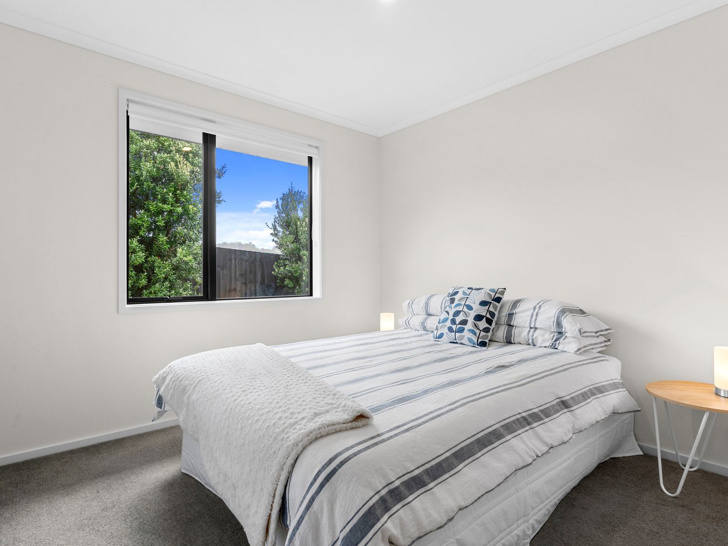 A bedroom with a bed striped bedding and two pillows next to a window and a small round wooden side table with a lamp at Mangawhai Maketu Mangawhai Holiday Home