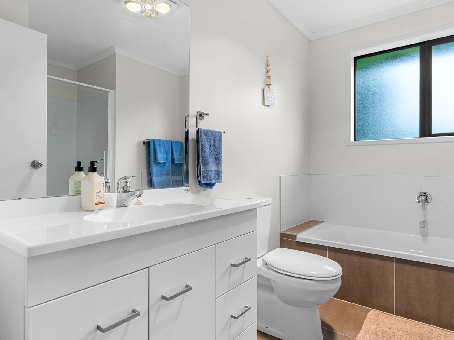A bathroom with a white vanity and sink, toilet, bathtub with brown tiles, blue towels, and a frosted window at Mangawhai Maketu Mangawhai