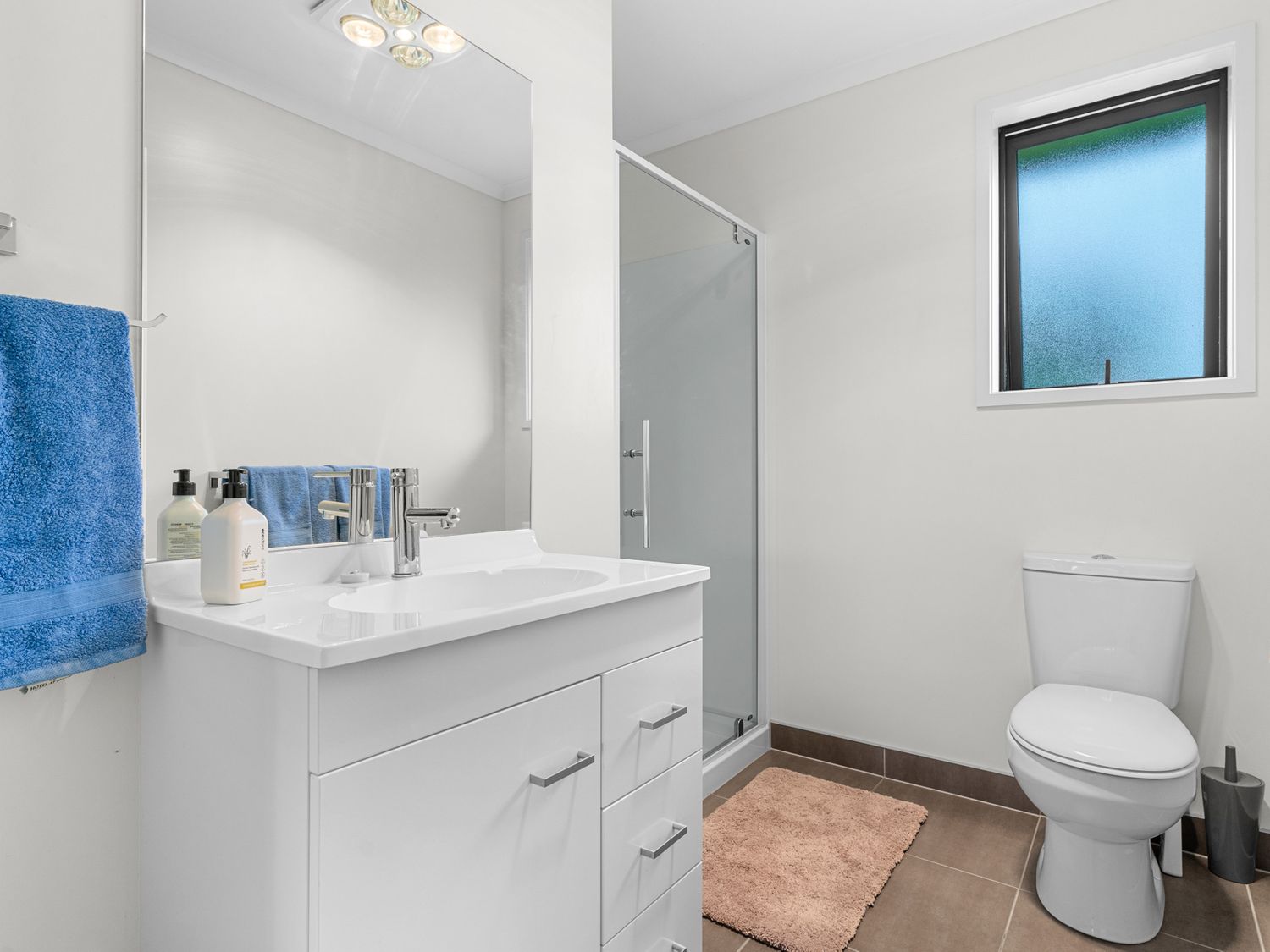 A bathroom with a white vanity and sink a mirror a glass shower door a toilet a window a blue towel and a beige rug at Mangawhai Maketu - Mangawhai Holiday Home