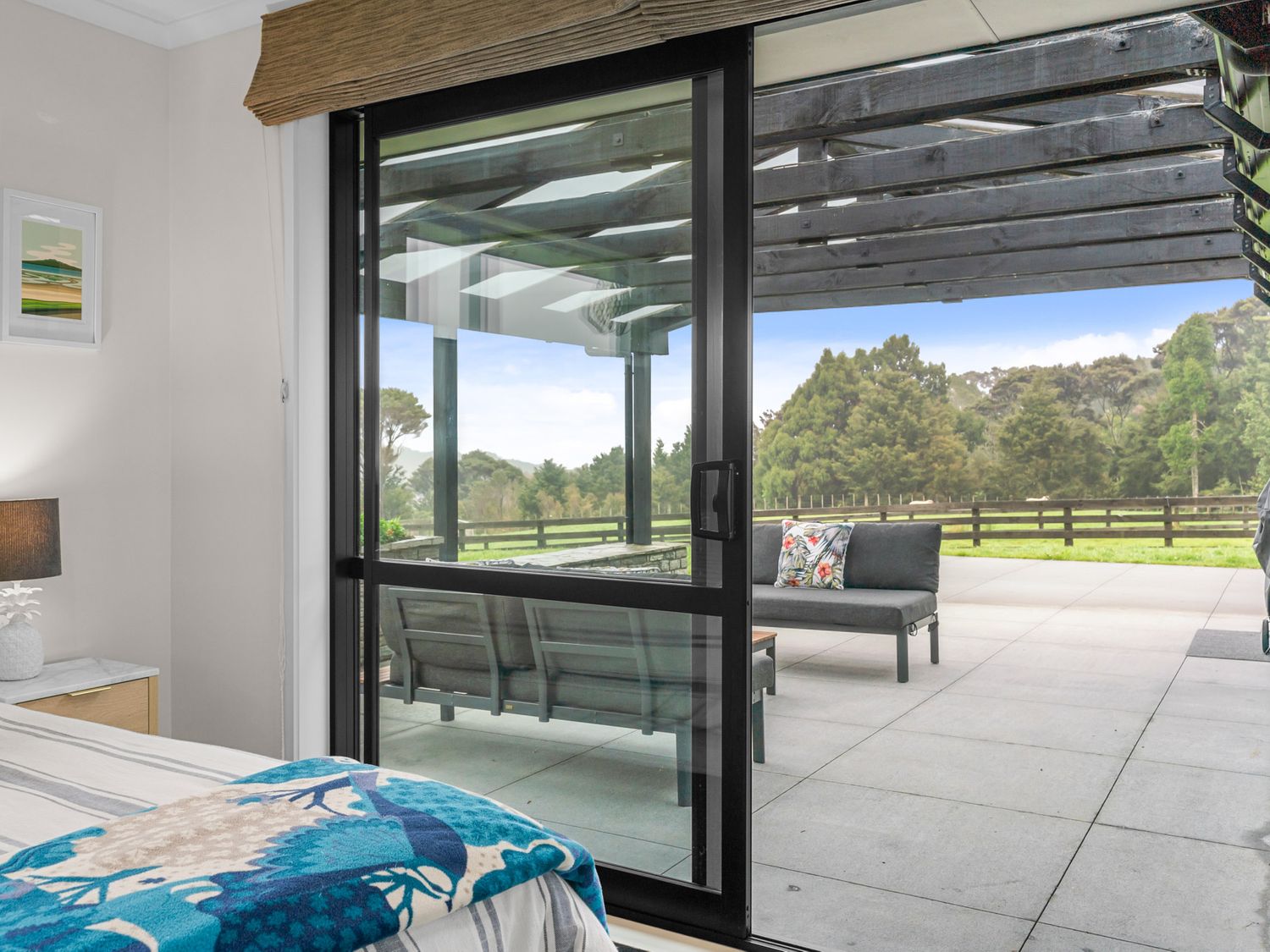 A bedroom with a bed and nightstand next to a sliding glass door overlooking an outdoor patio with seating and a view of trees at Mangawhai Maketu Mangawhai