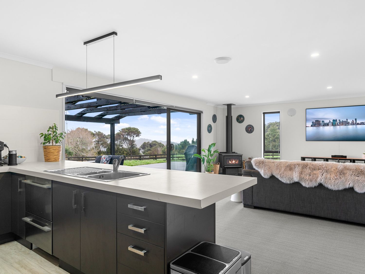 A kitchen island with sink and drawers overlooking a living room with a sofa and a wood stove at Mangawhai Maketu Mangawhai