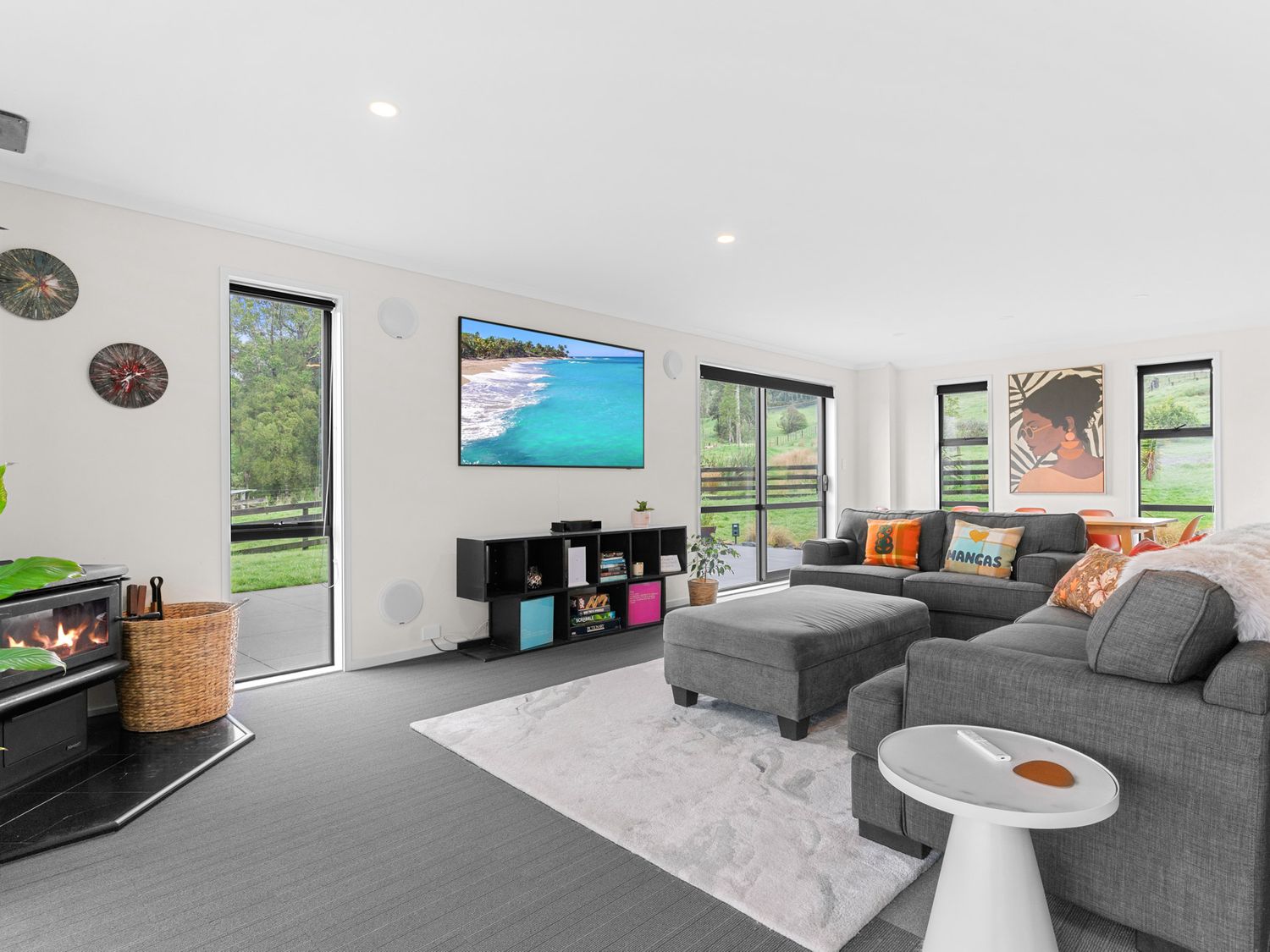 A living room with a gray sectional sofa and ottoman a white side table a TV on a black shelf two windows and a fireplace at Mangawhai Maketu Mangawhai