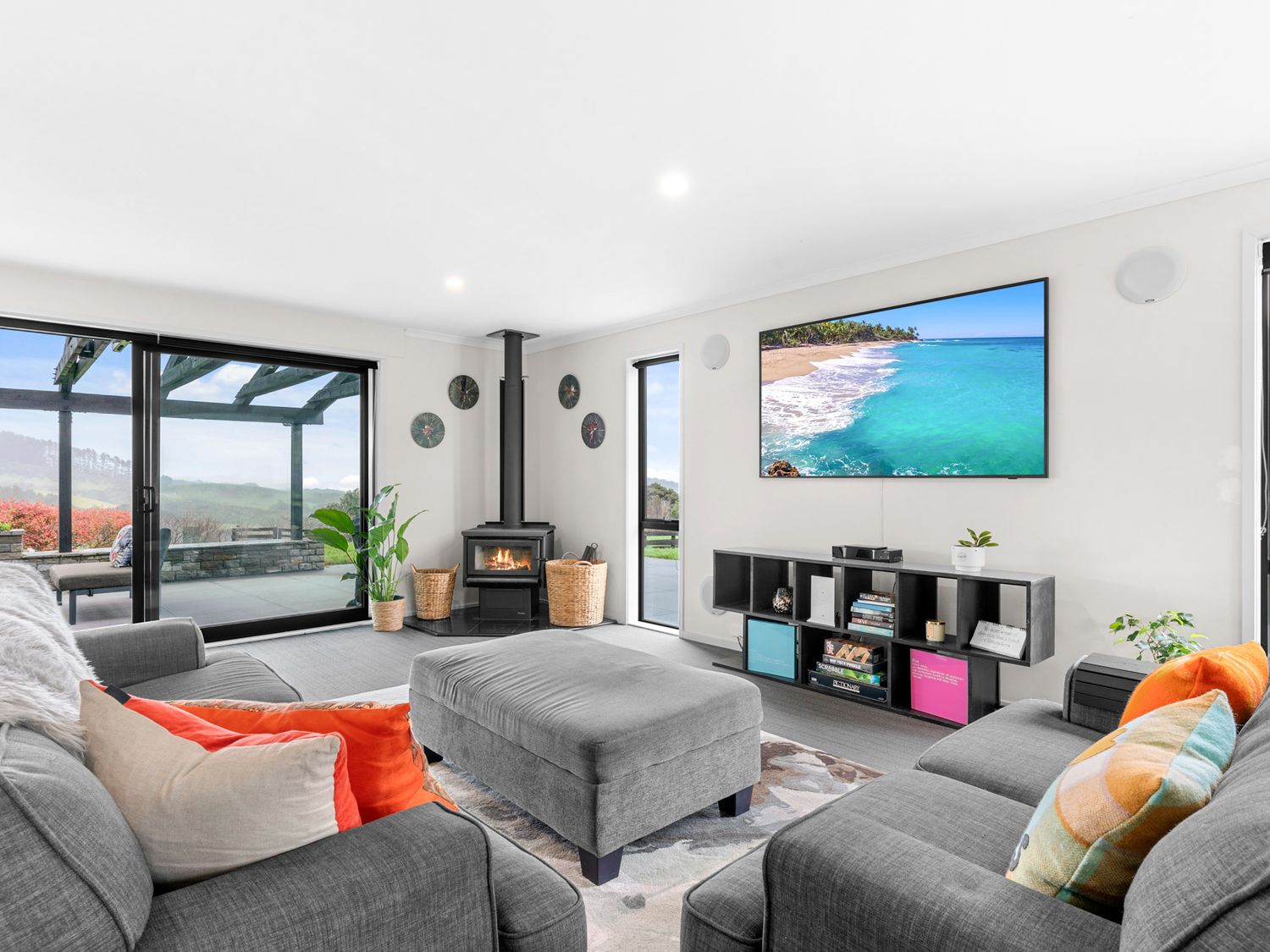 A living room with grey sofas a grey ottoman a wood stove and a wall-mounted tv at Mangawhai Maketu Mangawhai