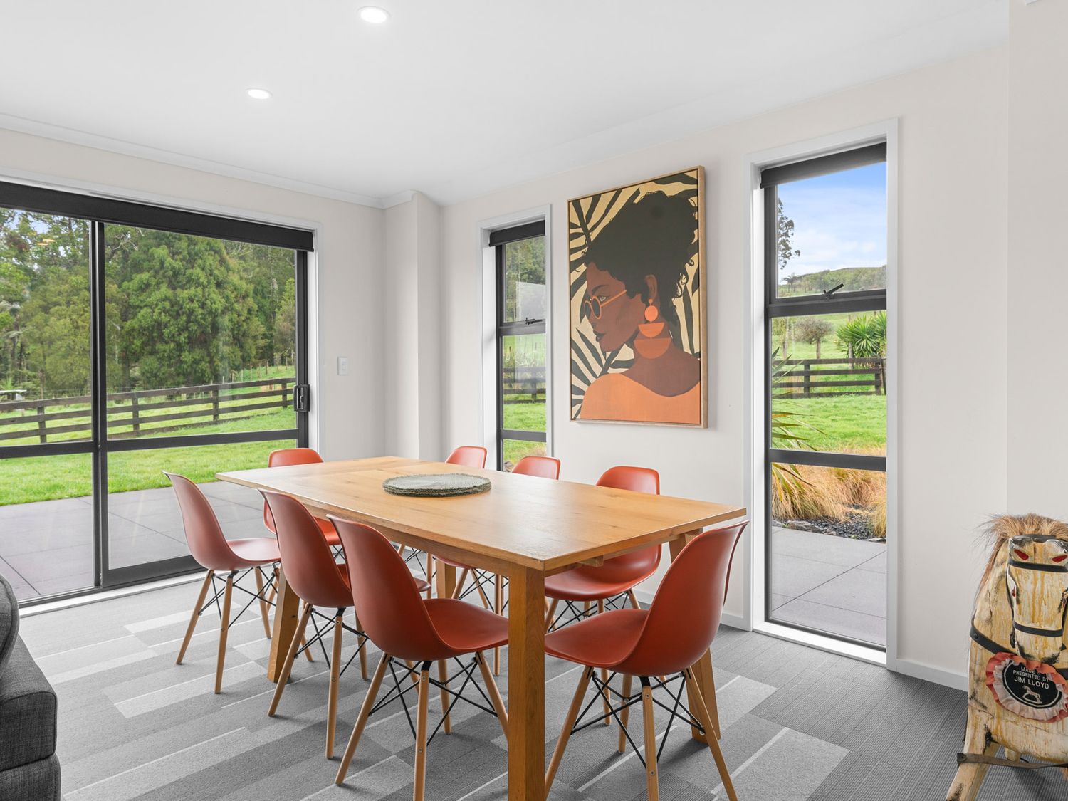 A dining room with a wooden table and eight orange chairs near sliding glass doors and windows with a painting on the wall at Mangawhai Maketu Mangawhai