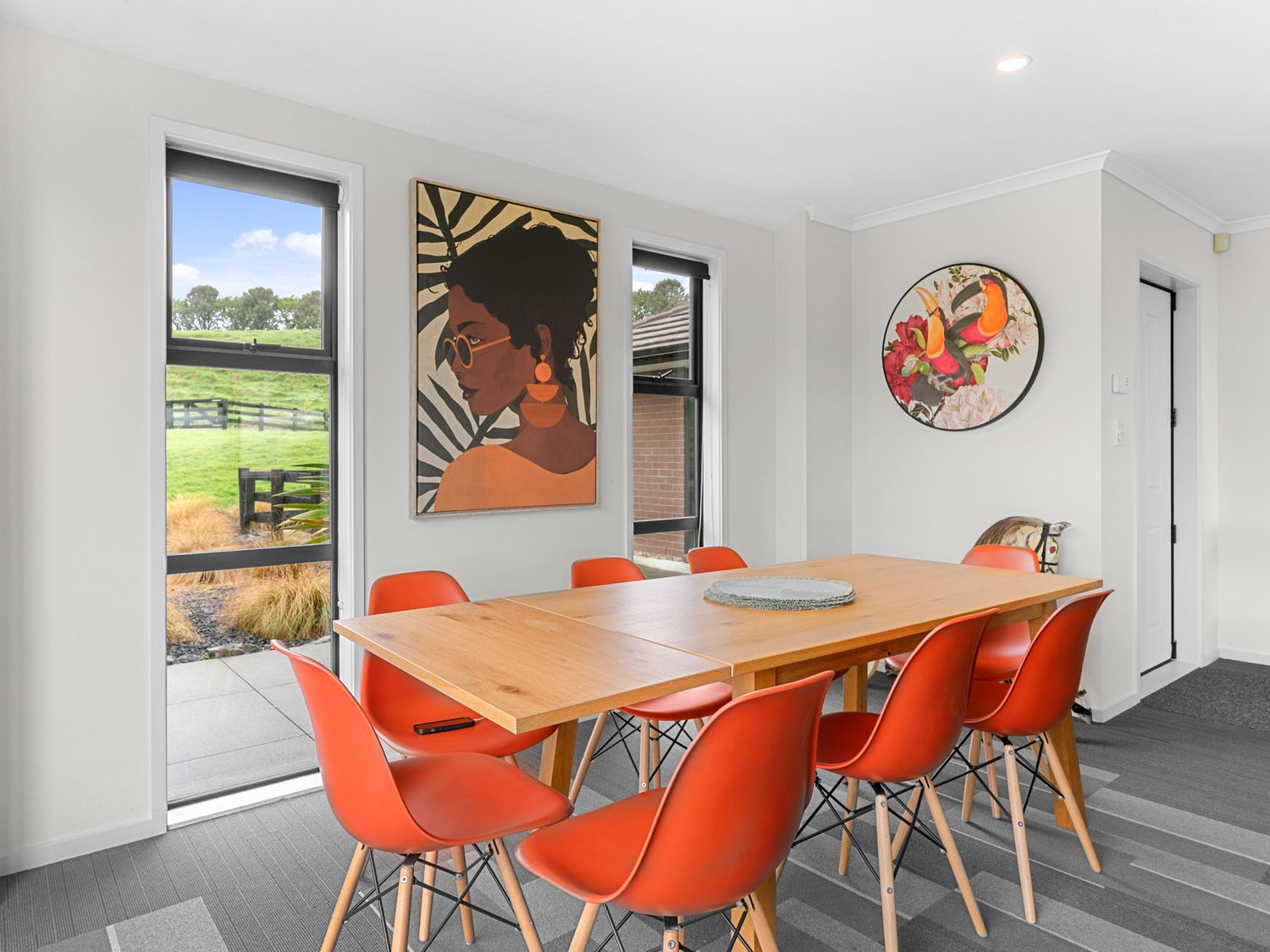 A dining room with a wooden table and eight orange chairs with artwork on the walls at Mangawhai Maketu Mangawhai