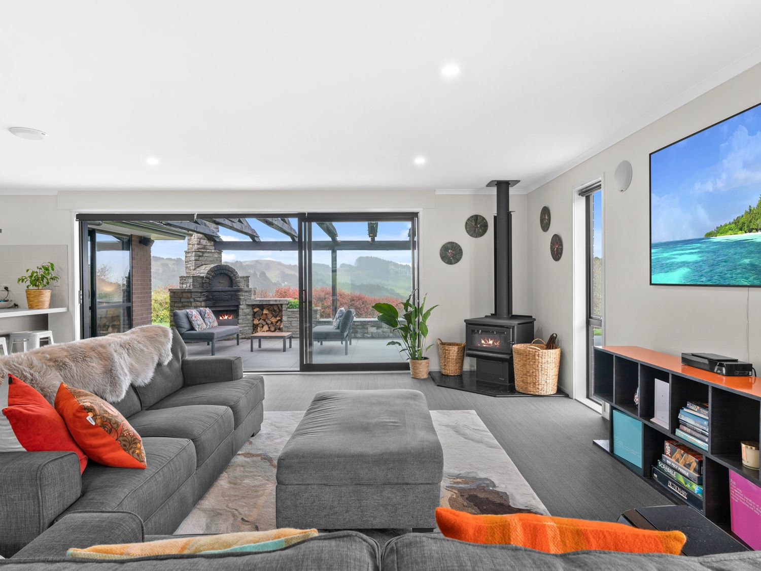 A living room with a sectional sofa and ottoman facing a wood stove and wall-mounted TV with sliding glass doors leading to an outdoor patio with a stone fireplace at Mangawhai Maketu in Mangawhai