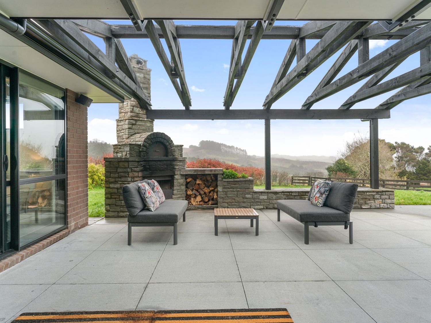 An outdoor patio with two gray sofas and a wooden coffee table under a wooden pergola with a stone fireplace and firewood storage at Mangawhai Maketu Mangawhai