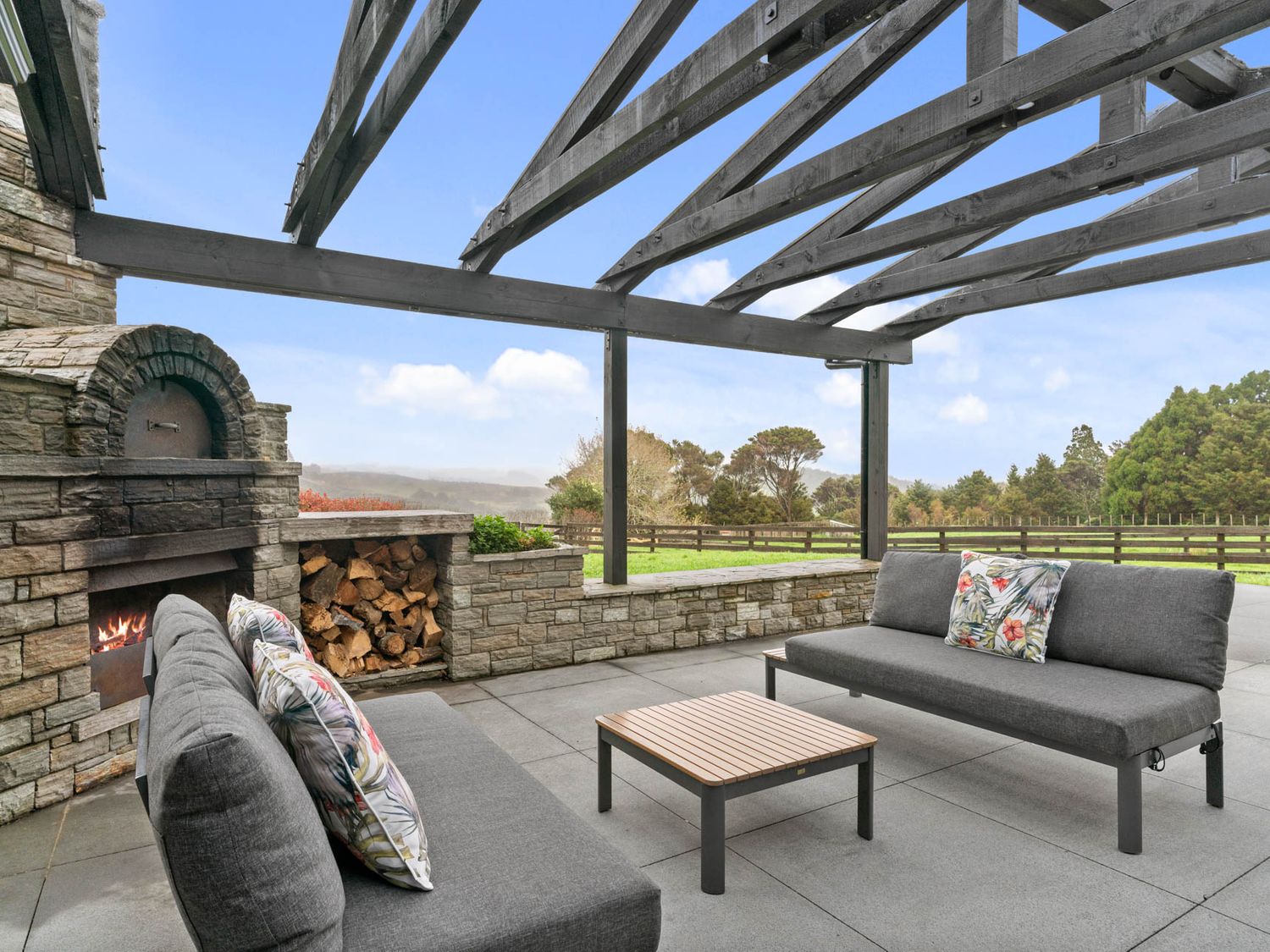 An outdoor seating area with two gray sofas, a wooden table, a stone fireplace with firewood, and a wooden pergola at Mangawhai Maketu - Mangawhai Holiday Home Mangawhai