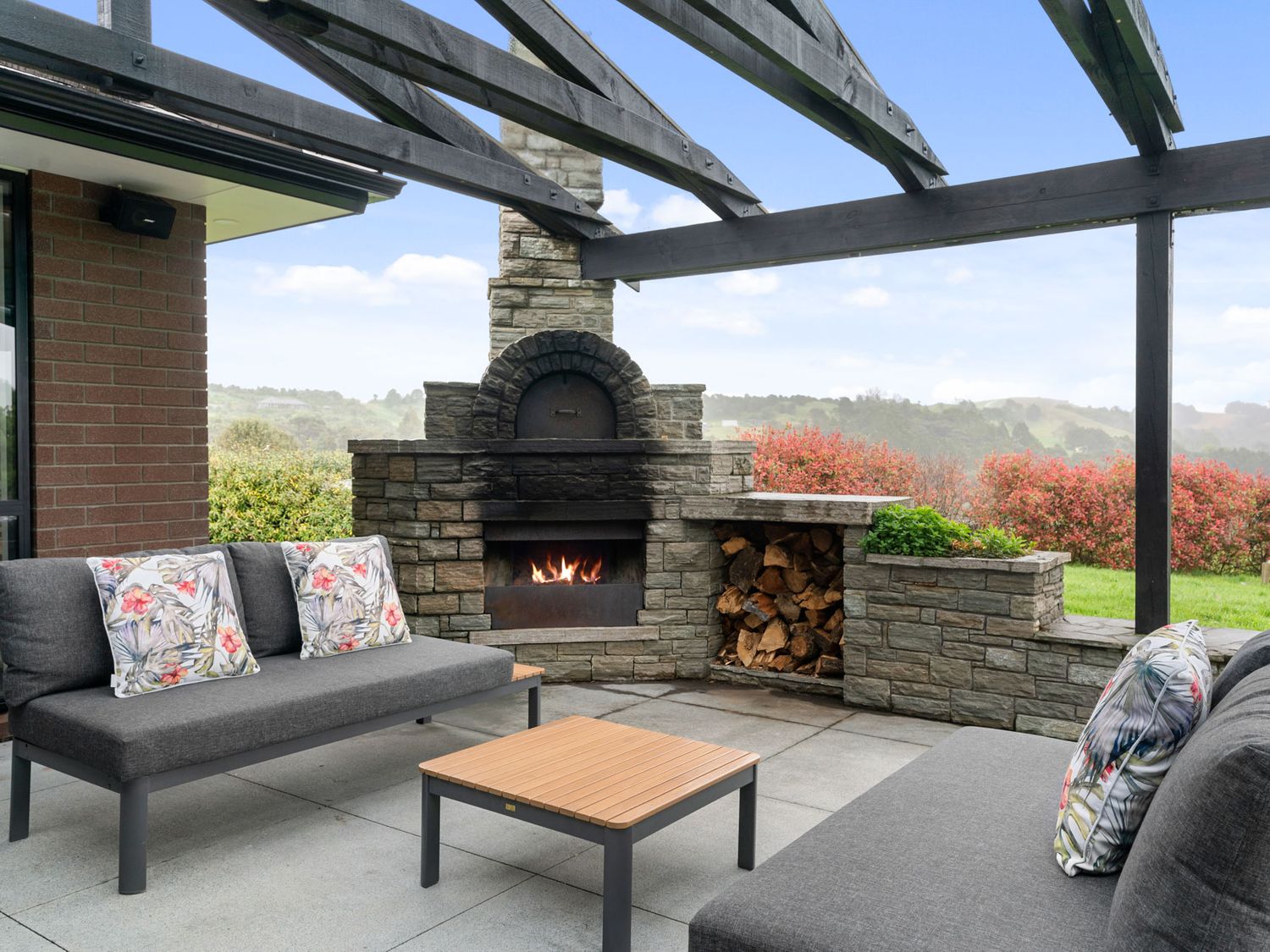 An outdoor patio with stone fireplace, two gray sofas with floral pillows, and a wooden coffee table at Mangawhai Maketu in Mangawhai