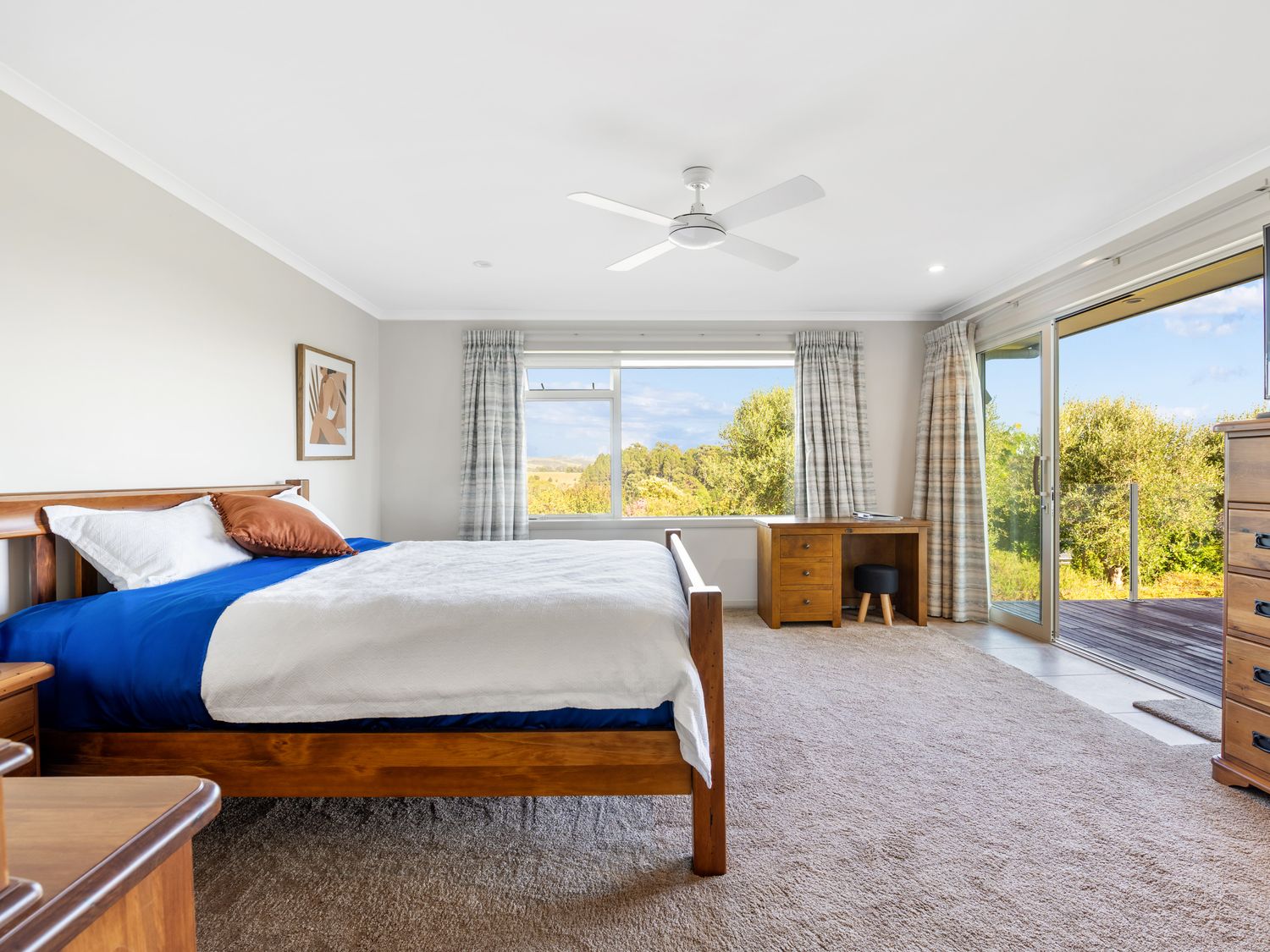 A bedroom with a wooden bed a desk a chest of drawers and glass doors to an outdoor deck at RidgeRaj - Kerikeri Holiday Home in Kerikeri