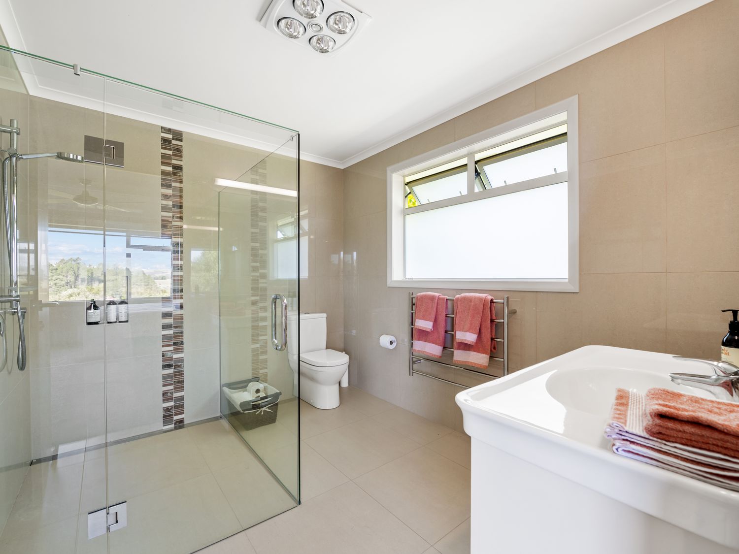 A bathroom with a glass shower enclosure a toilet a sink with folded towels and a window with pink towels hanging on a rack at RidgeRaj - Kerikeri Holiday Home in Kerikeri