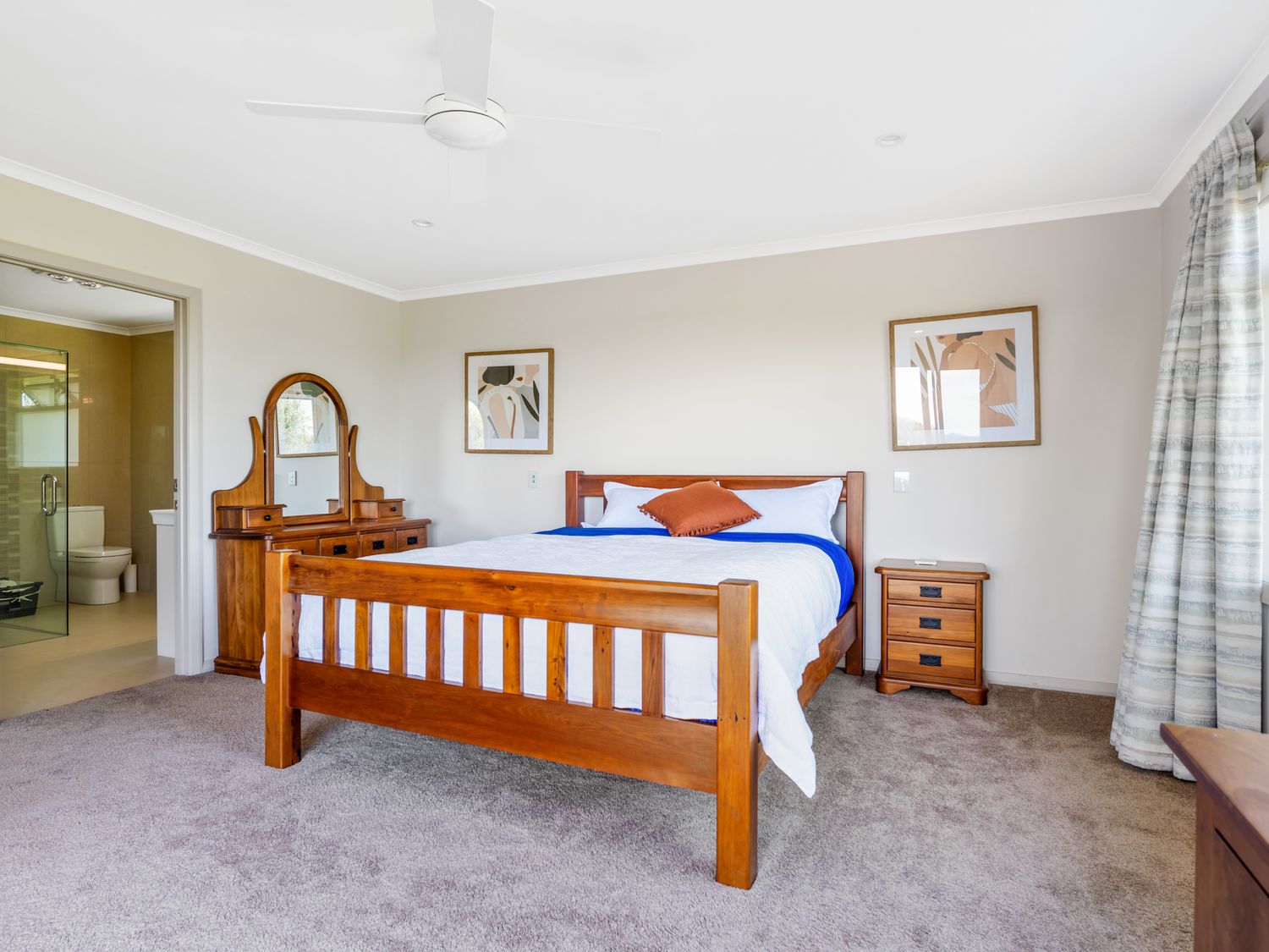 A bedroom with a wooden bed, bedside table, dresser with mirror, two framed pictures on the wall, and an ensuite bathroom at RidgeRaj - Kerikeri Holiday Home in Kerikeri