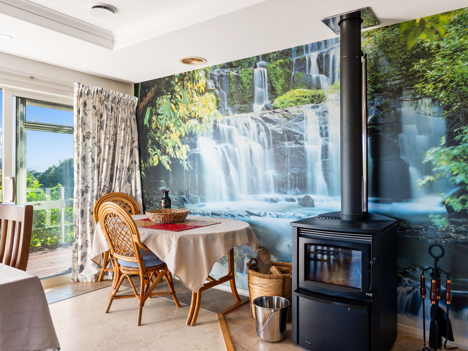 A dining area with two rattan chairs a table with a tablecloth and a black wood stove in front of a waterfall mural at RidgeRaj - Kerikeri Holiday Home in Kerikeri