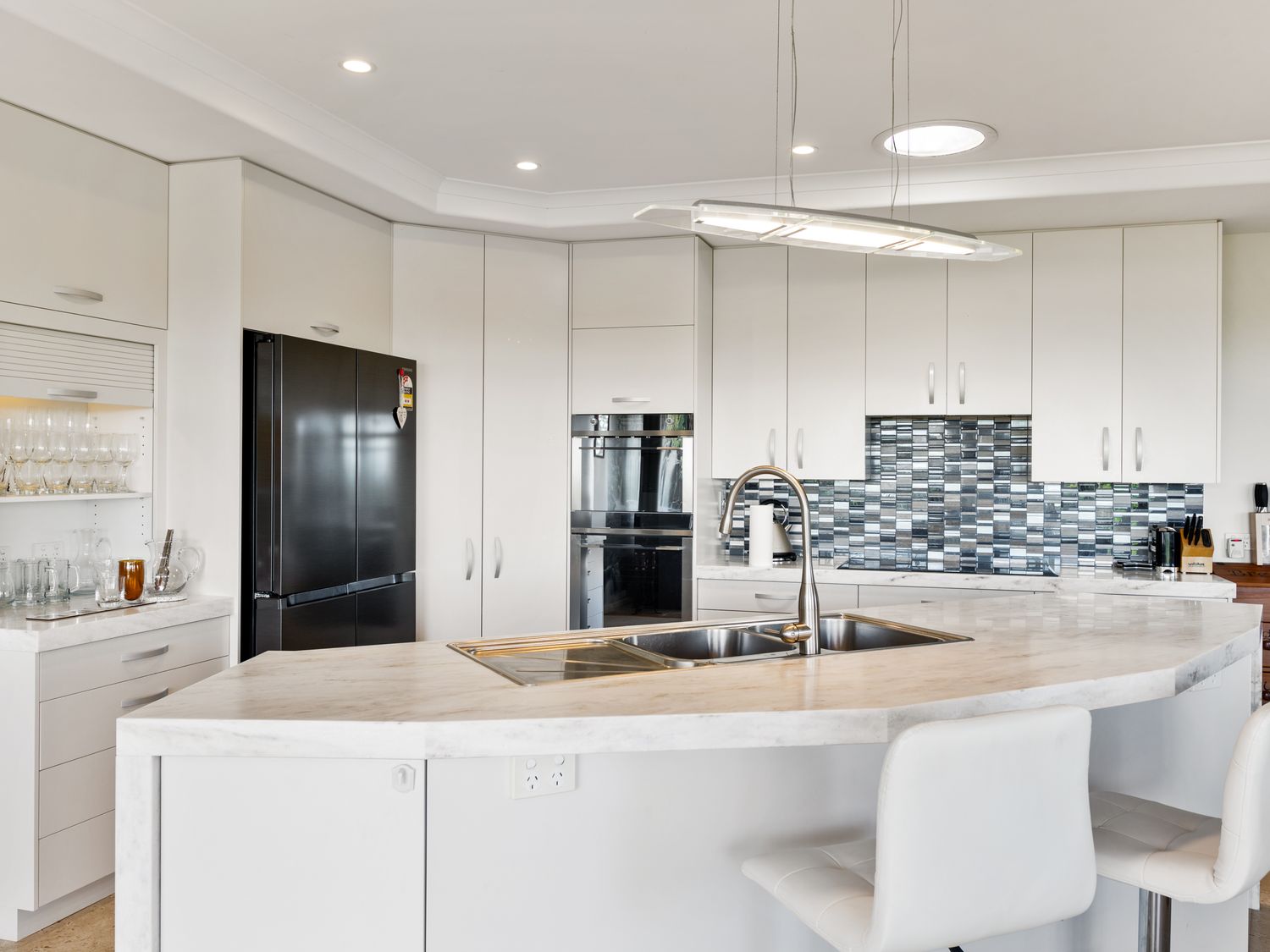 A kitchen with white cabinets a black refrigerator a double sink on a marble countertop and a mosaic tile backsplash at RidgeRaj - Kerikeri Holiday Home in Kerikeri