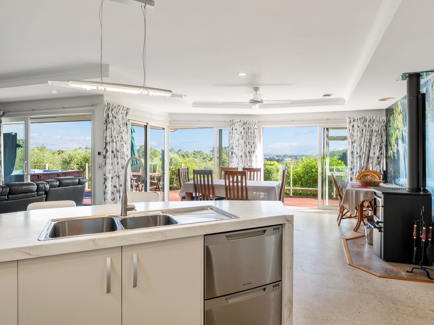 A kitchen with a sink and dishwasher overlooking a dining area with wooden chairs and tables at RidgeRaj Kerikeri Holiday Home Kerikeri