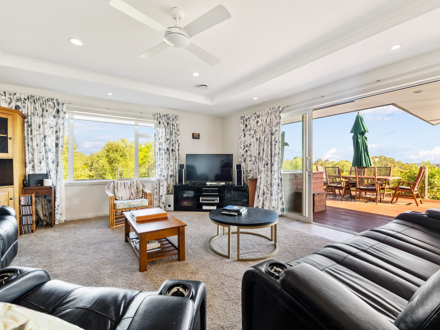 A living room with black leather sofas a wooden coffee table TV and sliding doors leading to a wooden deck with outdoor furniture at RidgeRaj - Kerikeri Holiday Home in Kerikeri