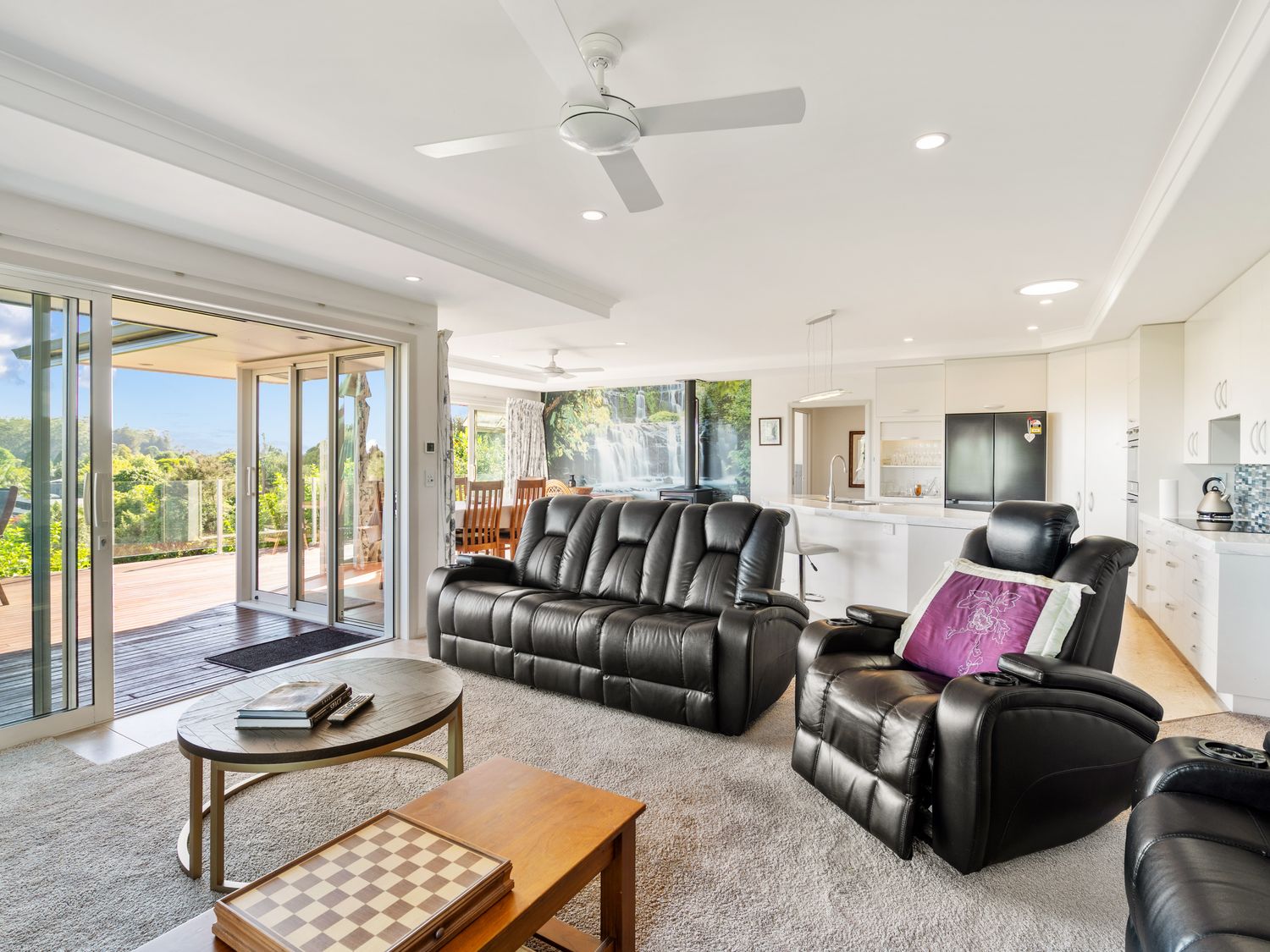 A living room with black leather sofas and recliner chairs next to a kitchen and dining area with sliding doors leading to an outdoor deck at RidgeRaj - Kerikeri Holiday Home in Kerikeri