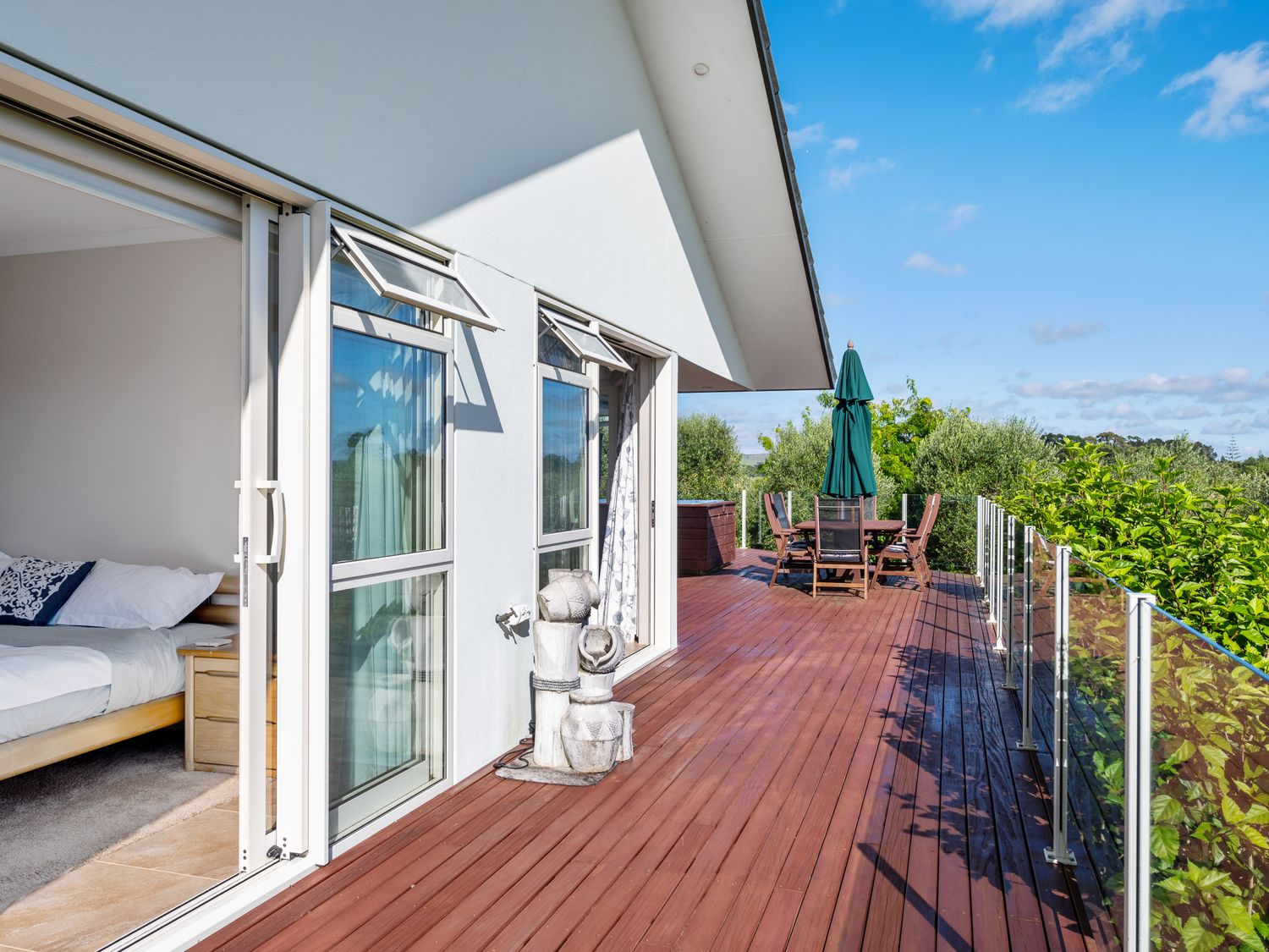 A wooden deck with outdoor dining furniture and umbrella next to a bedroom with open sliding doors at RidgeRaj - Kerikeri Holiday Home in Kerikeri