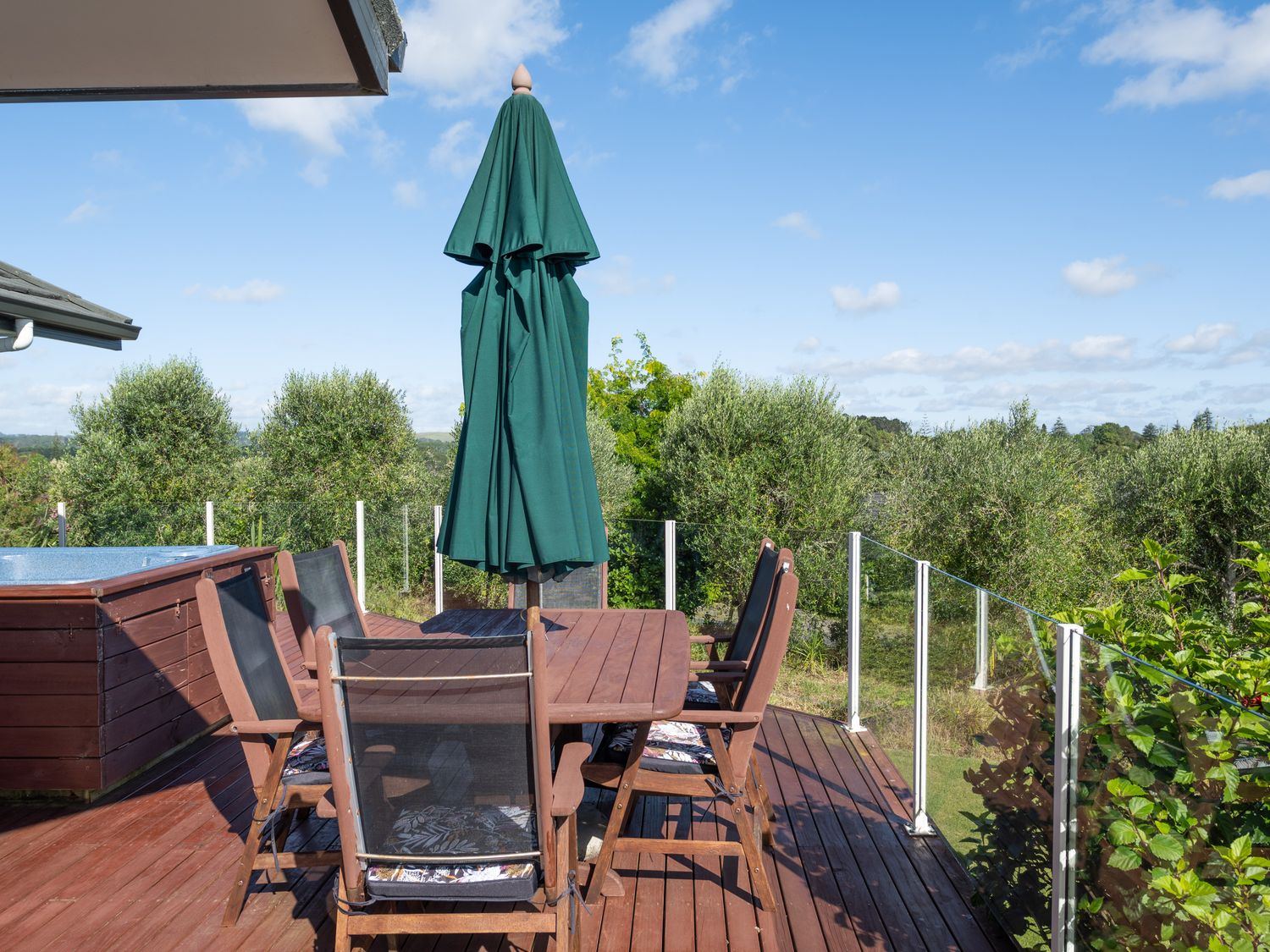 A wooden deck with a dining table six chairs and a closed green umbrella overlooking trees at RidgeRaj  Kerikeri Holiday Home in Kerikeri