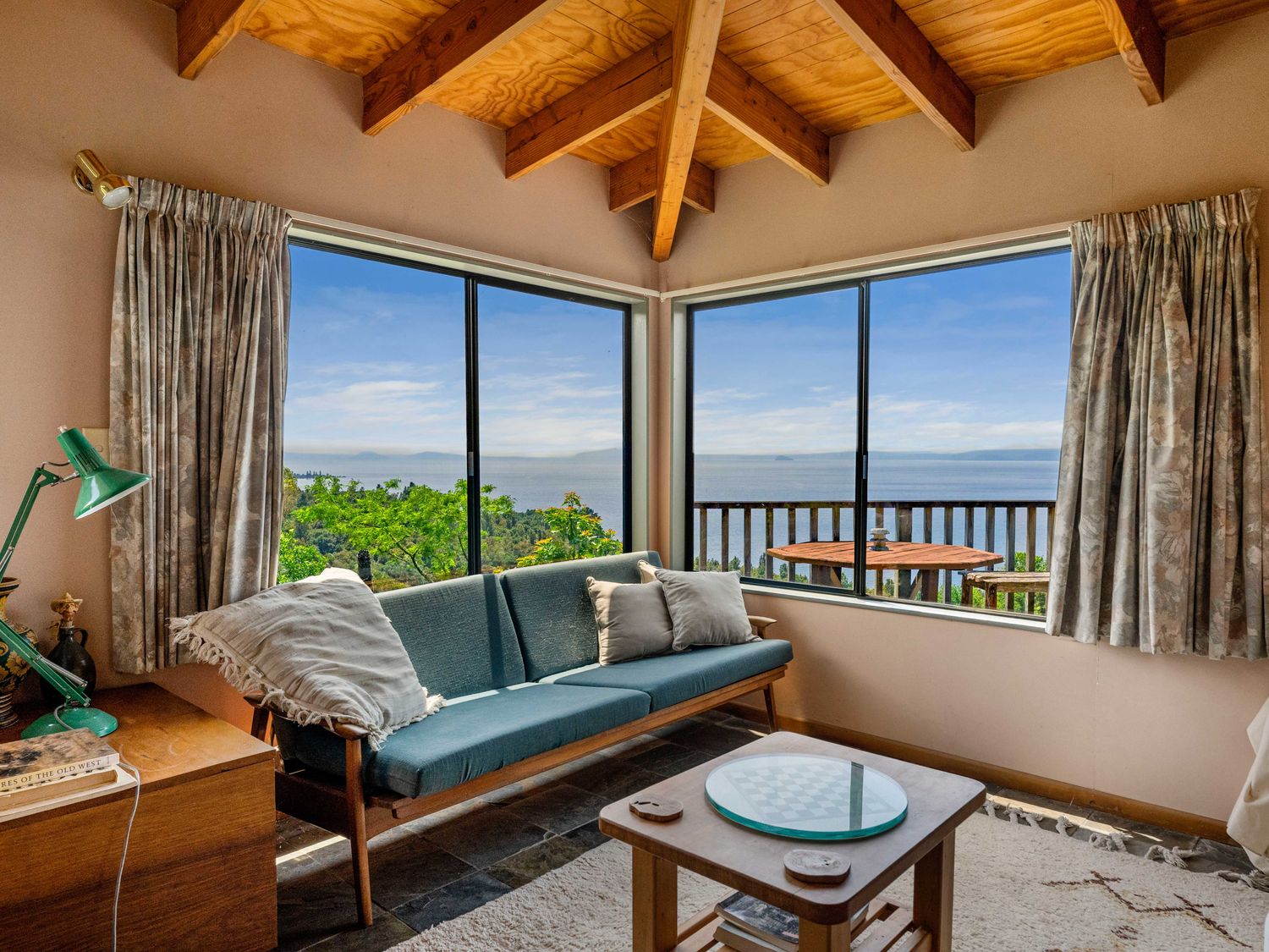 A living room with a blue sofa pillows a small wooden table with a chess board and large windows showing a water view at Pukawa Paradise - Pukawa Holiday Home in Pukawa Bay