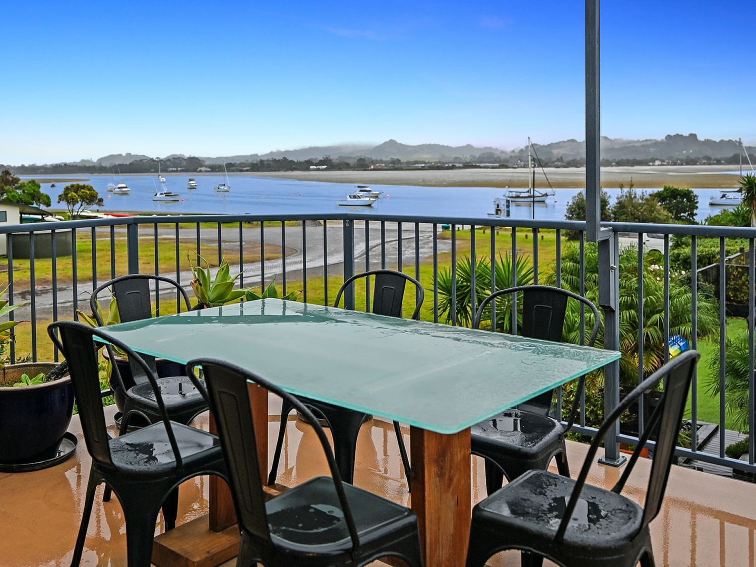 A balcony with a glass top table and six black metal chairs overlooking a river with boats and distant hills at Omaha Beach House in Omaha