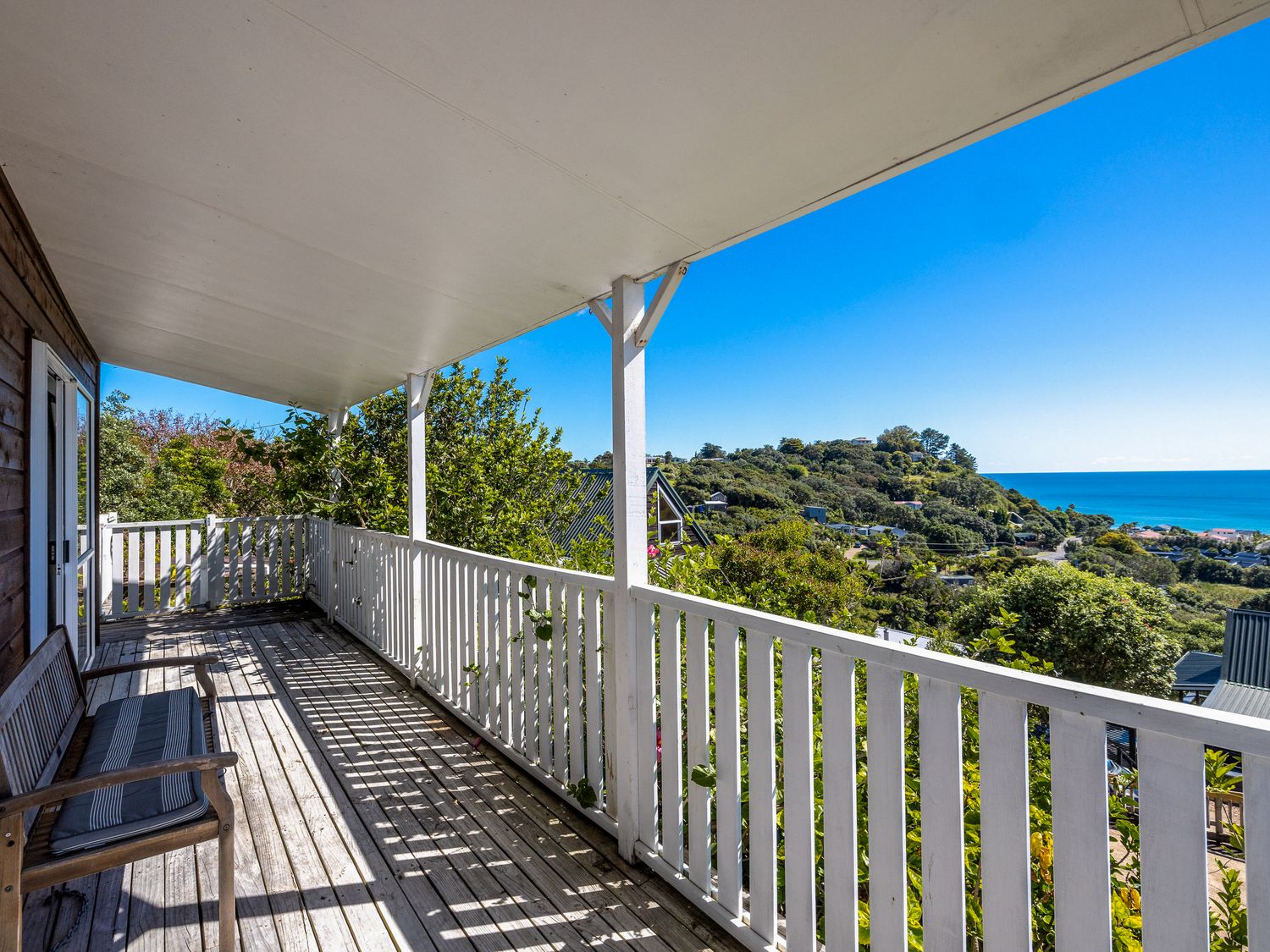A wooden balcony with white railing and a bench overlooking trees hills and the ocean at Oceanview Magic - Onetangi Holiday Home in Onetangi
