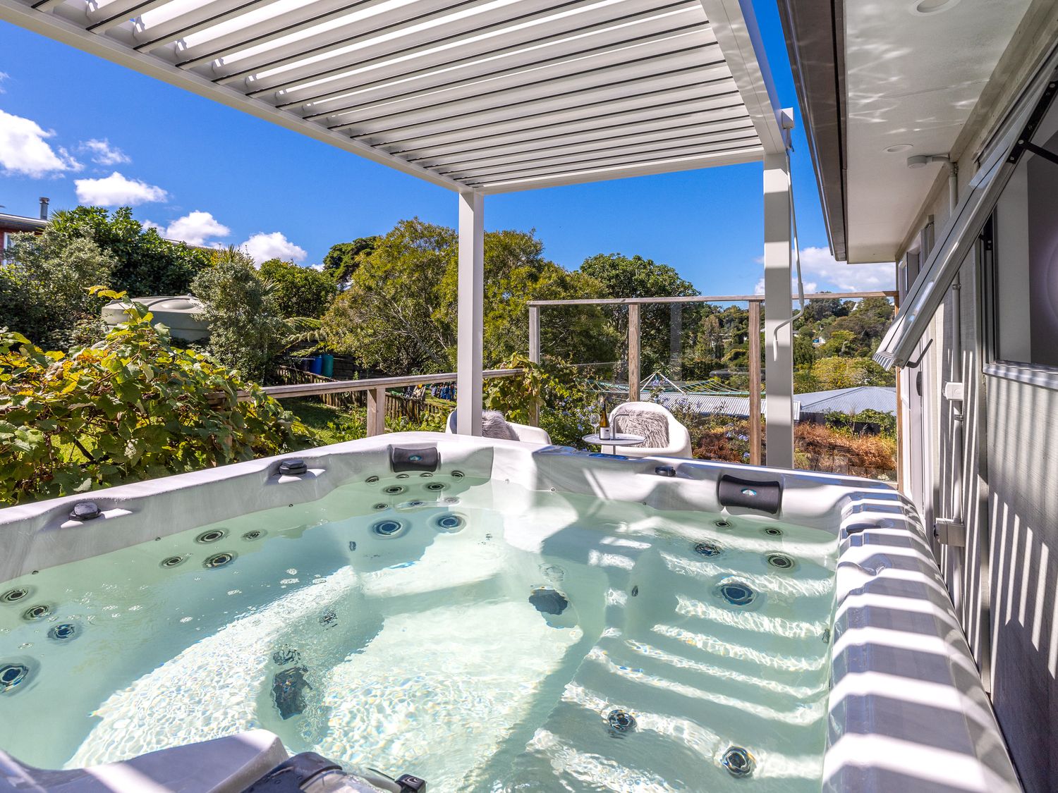 An outdoor hot tub on a deck with a pergola overhead and seating area with chairs and a table with a bottle at Surfdale Sunspot Spa Escape Waiheke in Surfdale