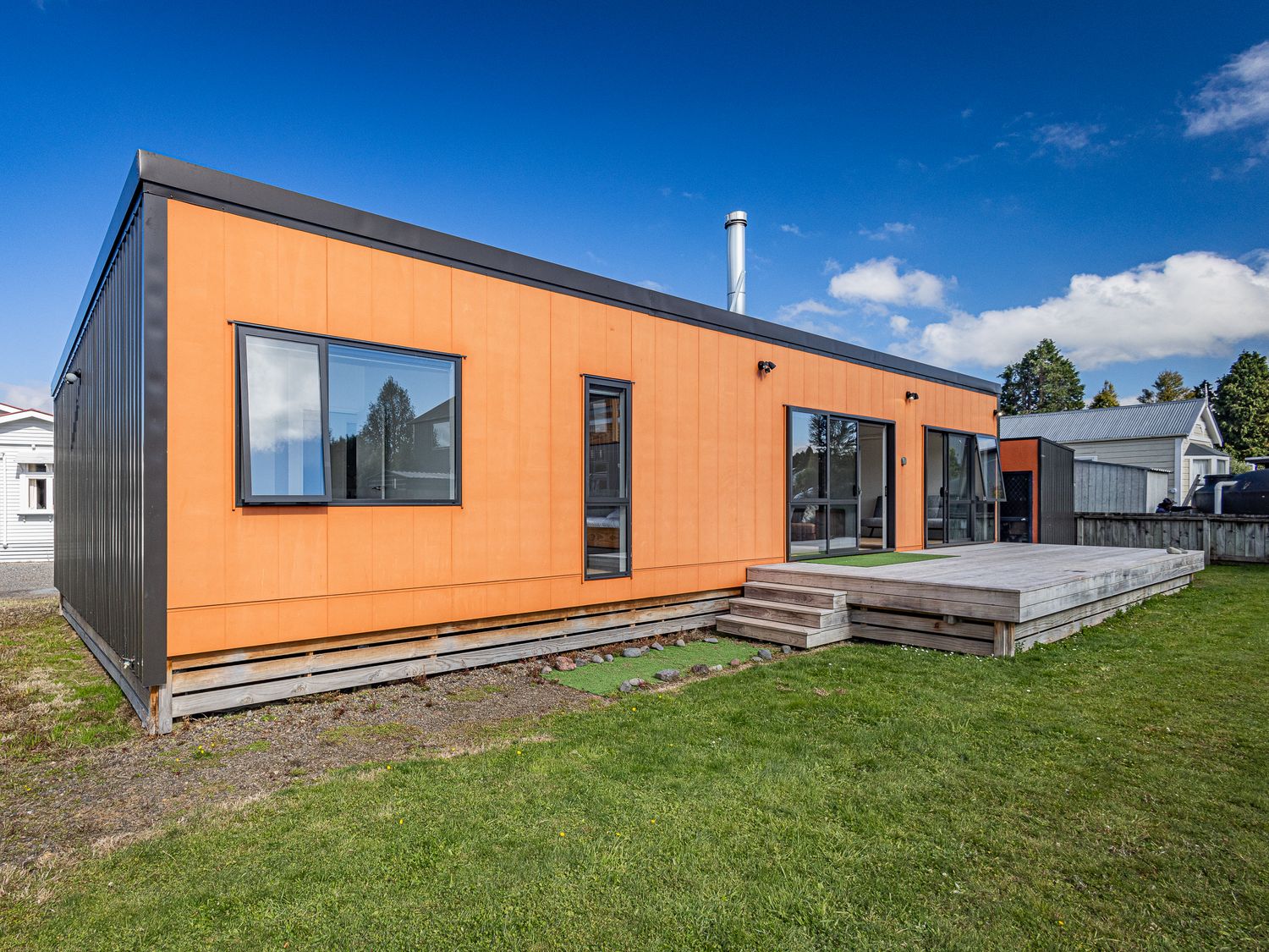 A modern orange and black single-story house with windows and a wooden deck in a grassy yard at Te Whare Retireti Hukarere - Rangataua Retreat