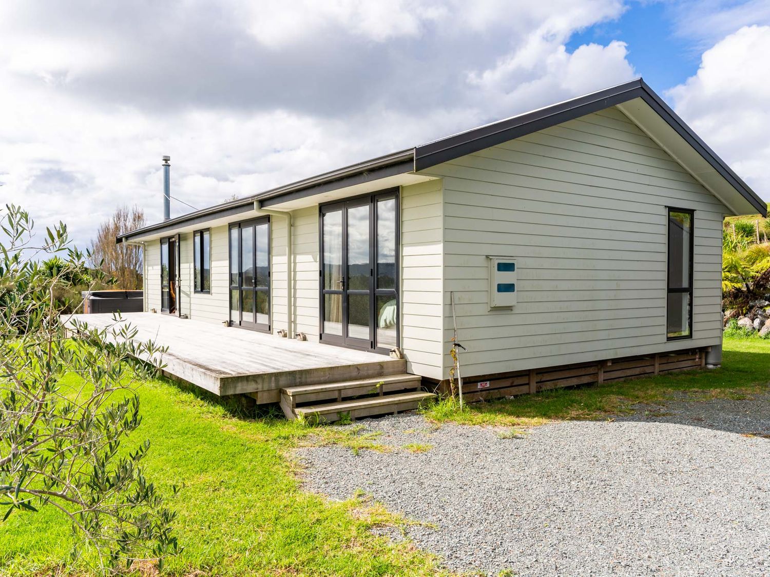 Exterior of a single story house with multiple glass doors and wooden deck next to grass and gravel area at The Olive Hut - Mangawhai Holiday Home in Mangawhai