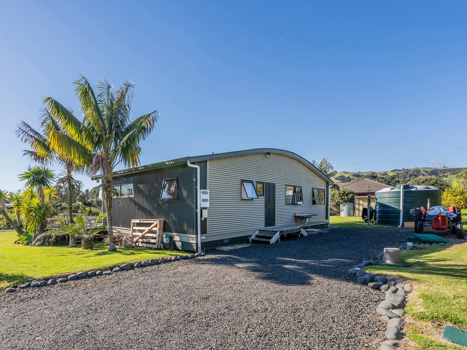 A corrugated metal building with small windows and a green door near palm trees and large water tanks at Wai Whare -  Kuaotunu Holiday Home in Kuaotunu