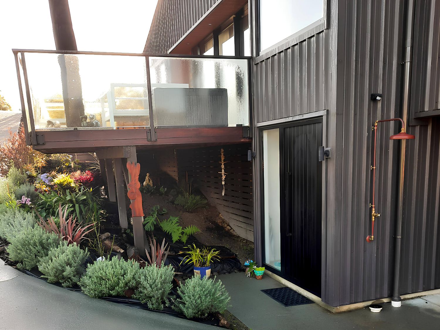 An exterior view of a modern building with a black door outdoor shower balcony with glass railing and garden plants at Coastal Serenity Langs Beach Holiday Home Waipu