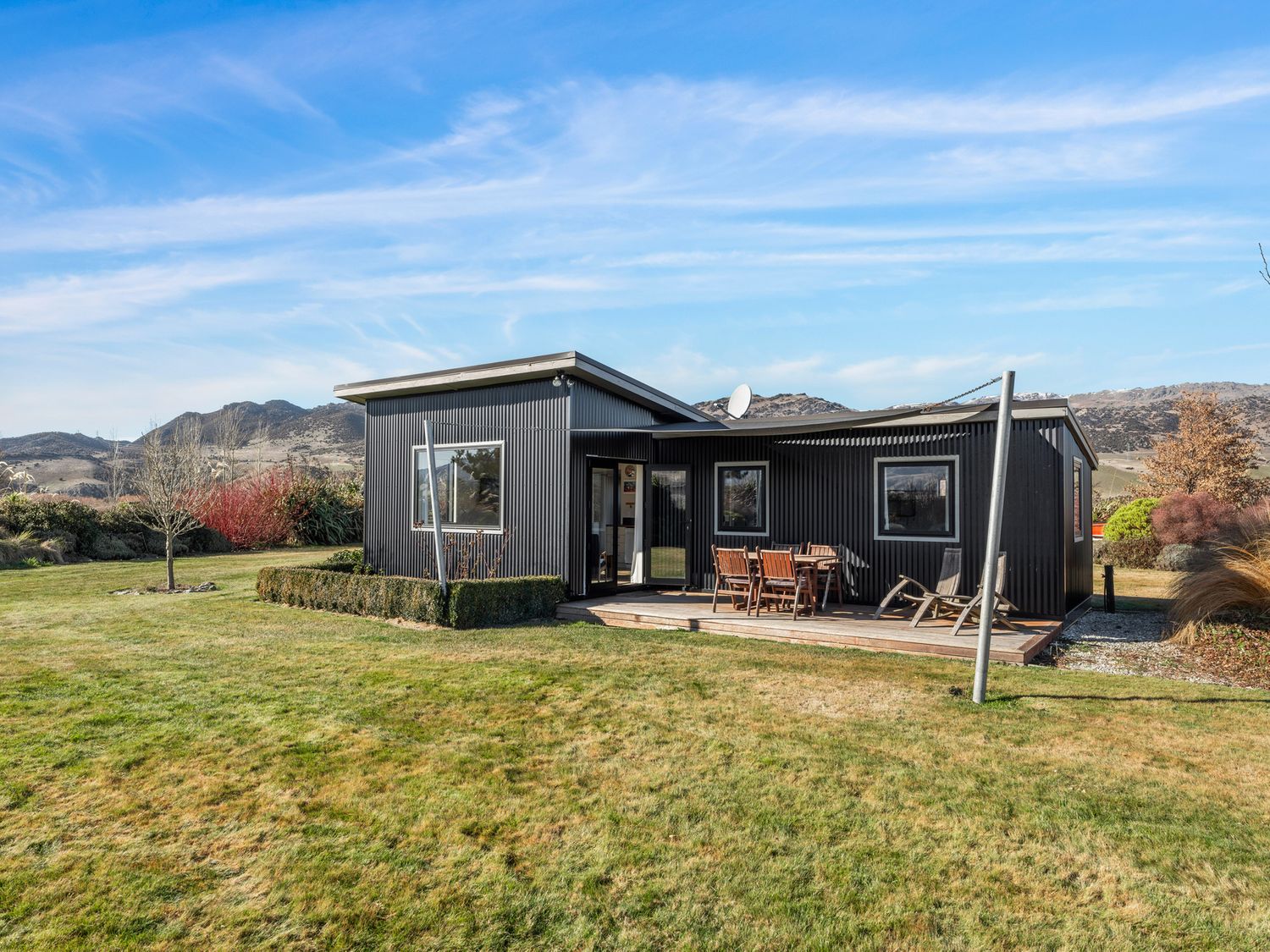 A black corrugated metal house with a wooden deck and outdoor dining furniture on a grassy yard with mountains in the background at Olive Cottage - Cromwell Holiday Home in Cromwell