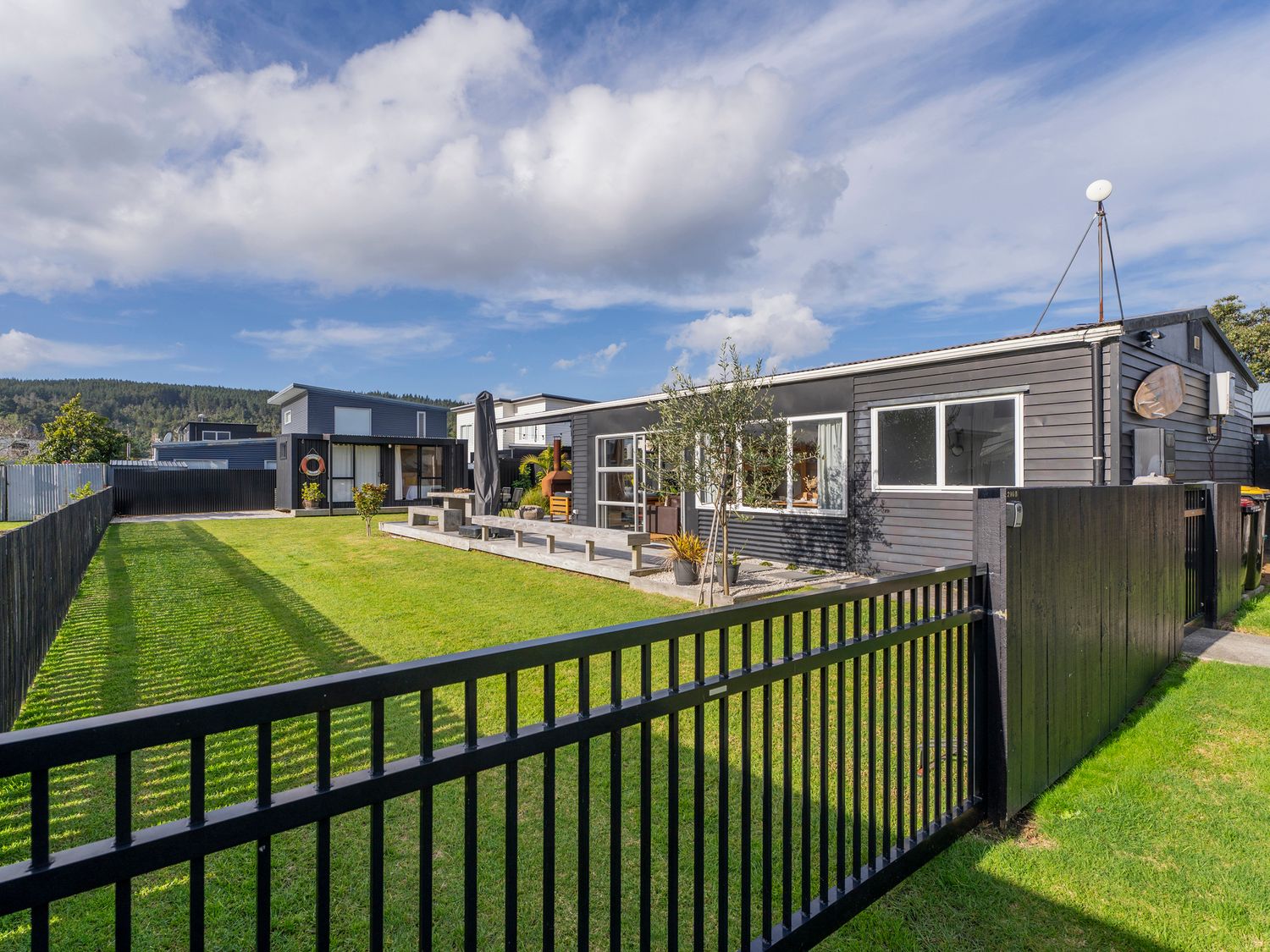 A black fenced yard with a grass lawn and a modern gray house with large windows at Sunny Hideaway  Whangamata