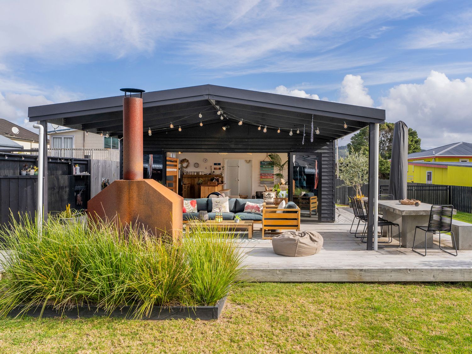 An outdoor patio area with a covered seating space a fireplace plants dining table and chairs at Sunny Hideaway - Whangamata Holiday Home in Whangamata