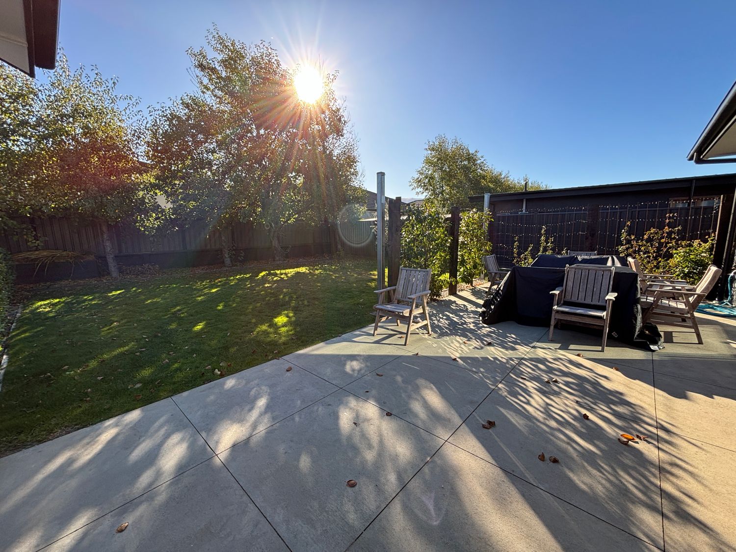 A backyard with a concrete patio, wooden chairs, grass, trees, and a fence at Pedal and Play - Cromwell Holiday Home in Cromwell