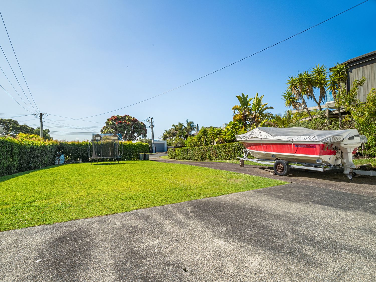 A driveway with a covered boat and a grassy yard with a trampoline surrounded by hedges at Unwind at Tindalls Bay - Whangaparaoa Apartment in Tindalls Bay