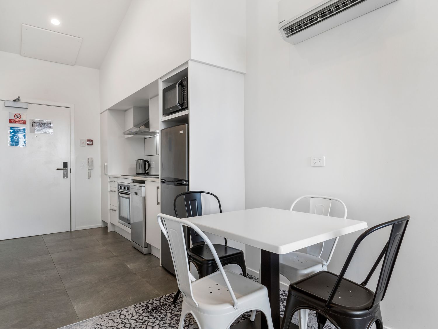A kitchen area with a white door a stove a dishwasher a refrigerator a microwave and a dining table with black and white chairs at Layard Apartments 531 - West Auckland Apartment in Avondale