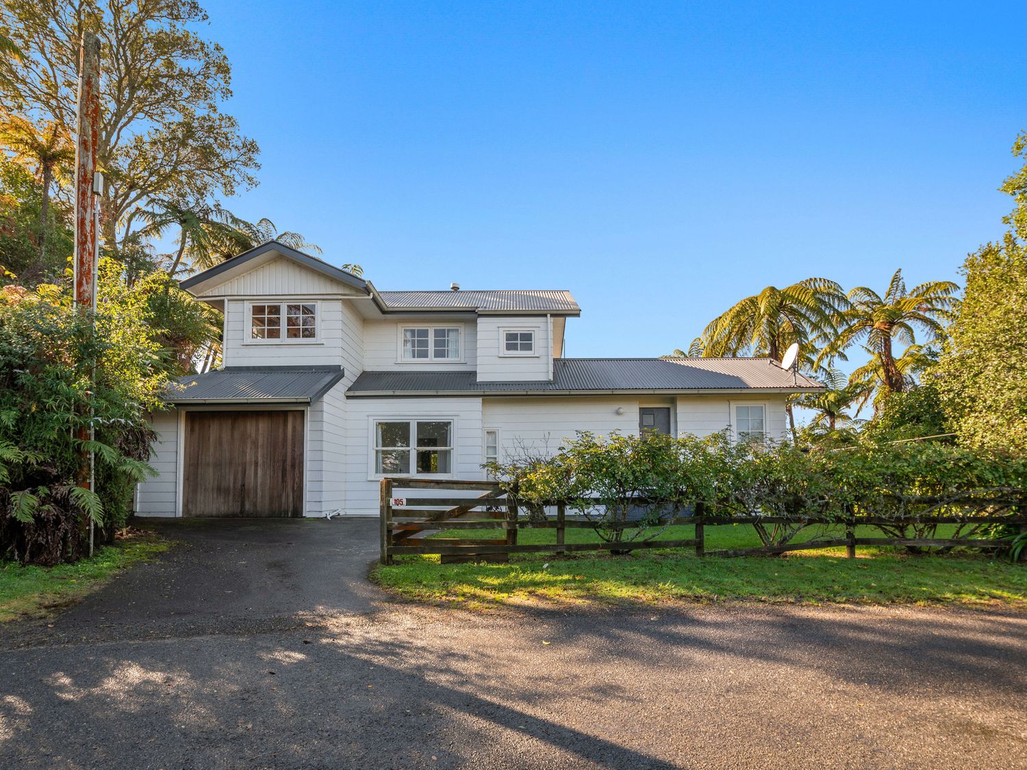 A white two-story house with a garage next to the driveway and a wooden fence surrounded by trees at Okere Retreat - Lake Rotoiti Getaway