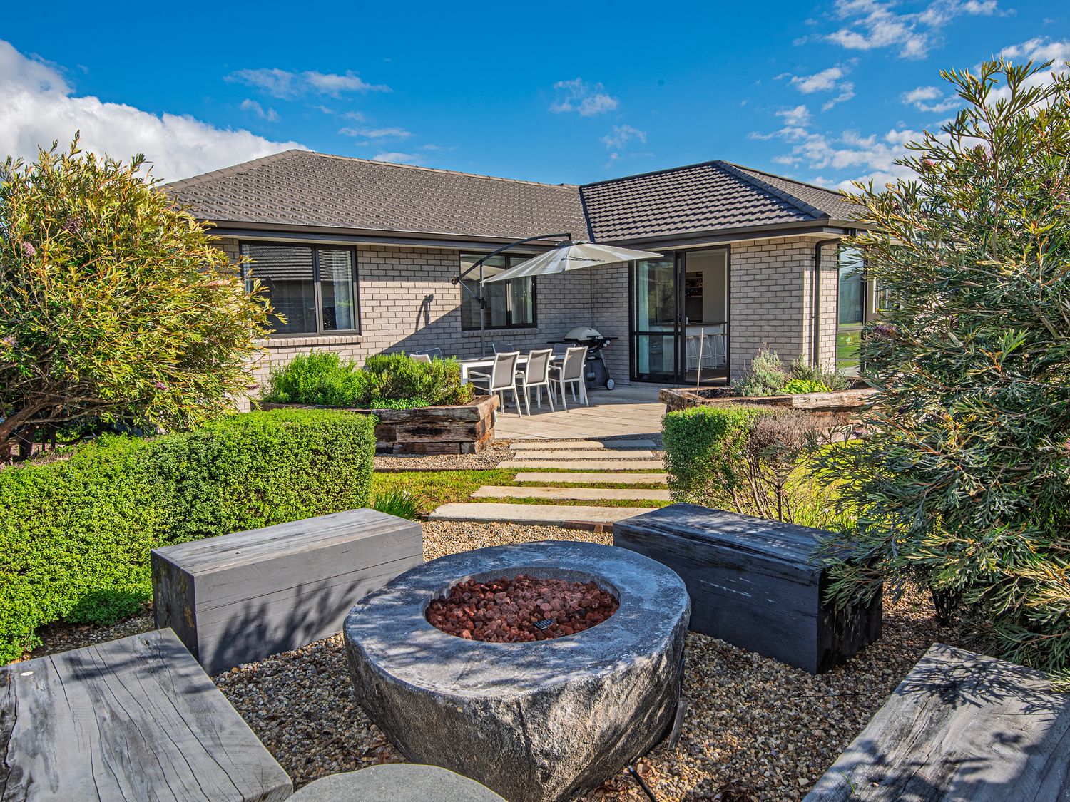 An outdoor seating area with benches and a firepit in a garden with bushes and stone steps leading to a patio with a dining table and umbrella at Bennett's Bach - Waipu Holiday Home in Waipu