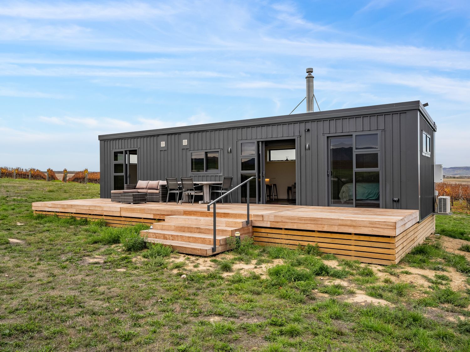 A modern dark gray modular home with wooden deck and outdoor furniture on grass at Alex Vineyard Alexandra Holiday Home Alexandra