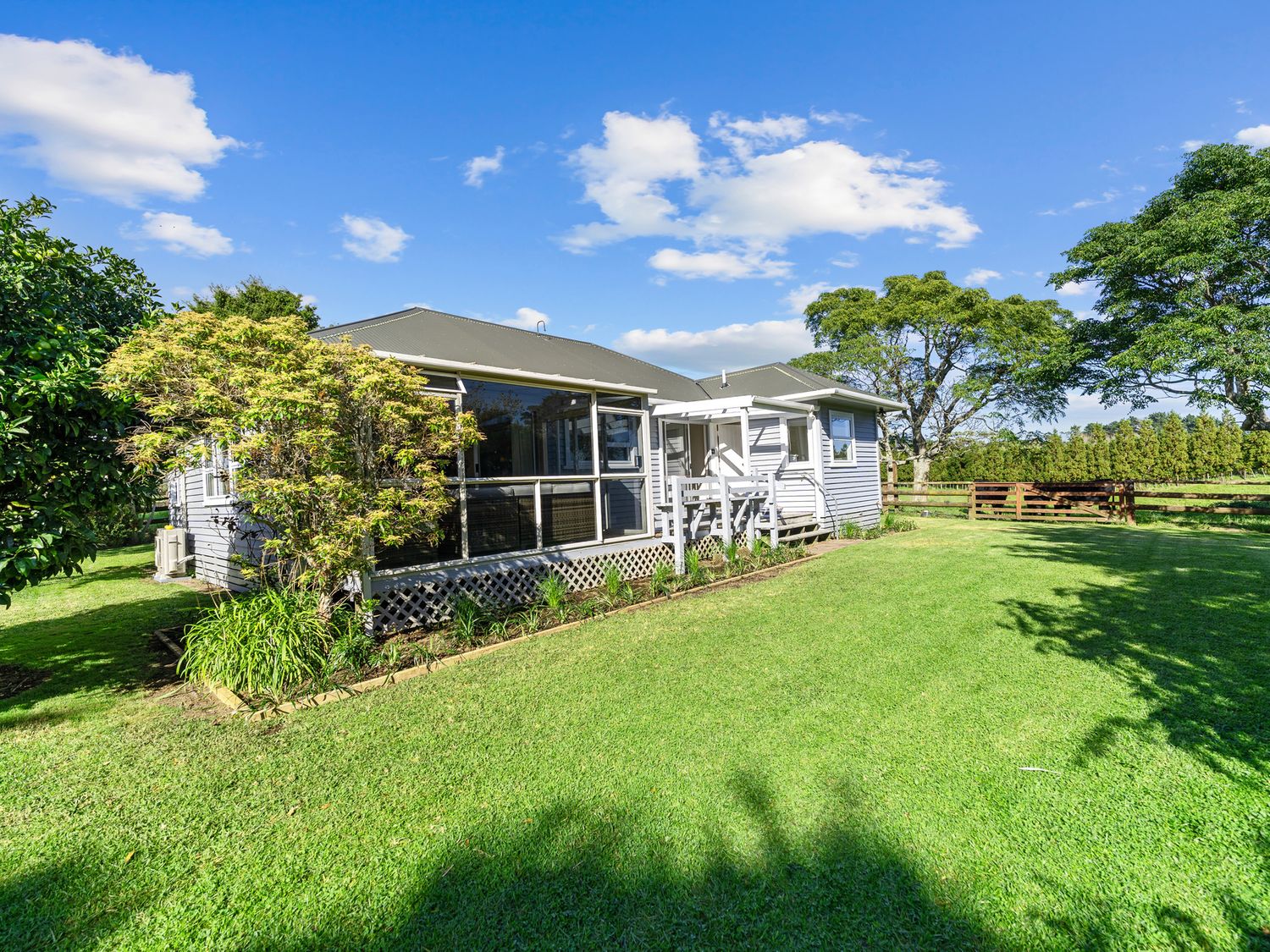 An exterior view of a single-story house with a porch and a lawn surrounded by trees at Bell Cottage - Oakura Holiday Home in Oakura