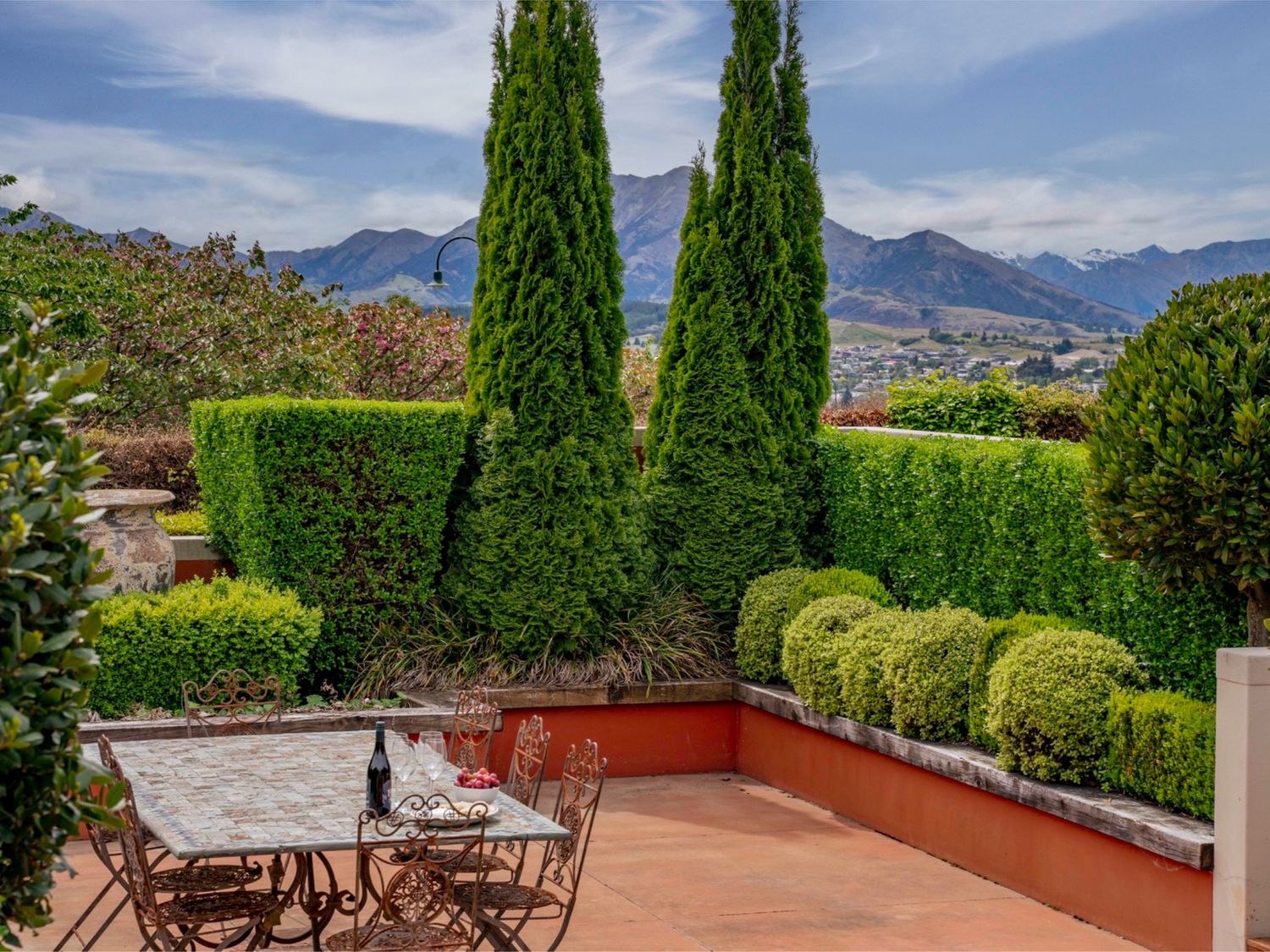 An outdoor patio area with a table and chairs surrounded by neatly trimmed hedges and trees with mountains in the background at Elderberry Wine - Wanaka Holiday Home in Wanaka