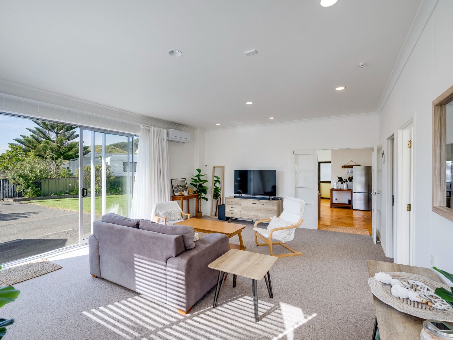 A living room with a sofa, two chairs, a coffee table, a television on a wooden stand, and sliding glass doors at Mahia Bliss - Surf & Serenity Holiday Home Mahia Peninsula