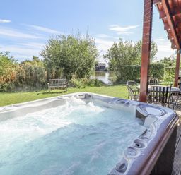 An outdoor hot tub with bubbling water next to wooden deck with table and chairs and greenery at Jemima Lodge in Tattershall